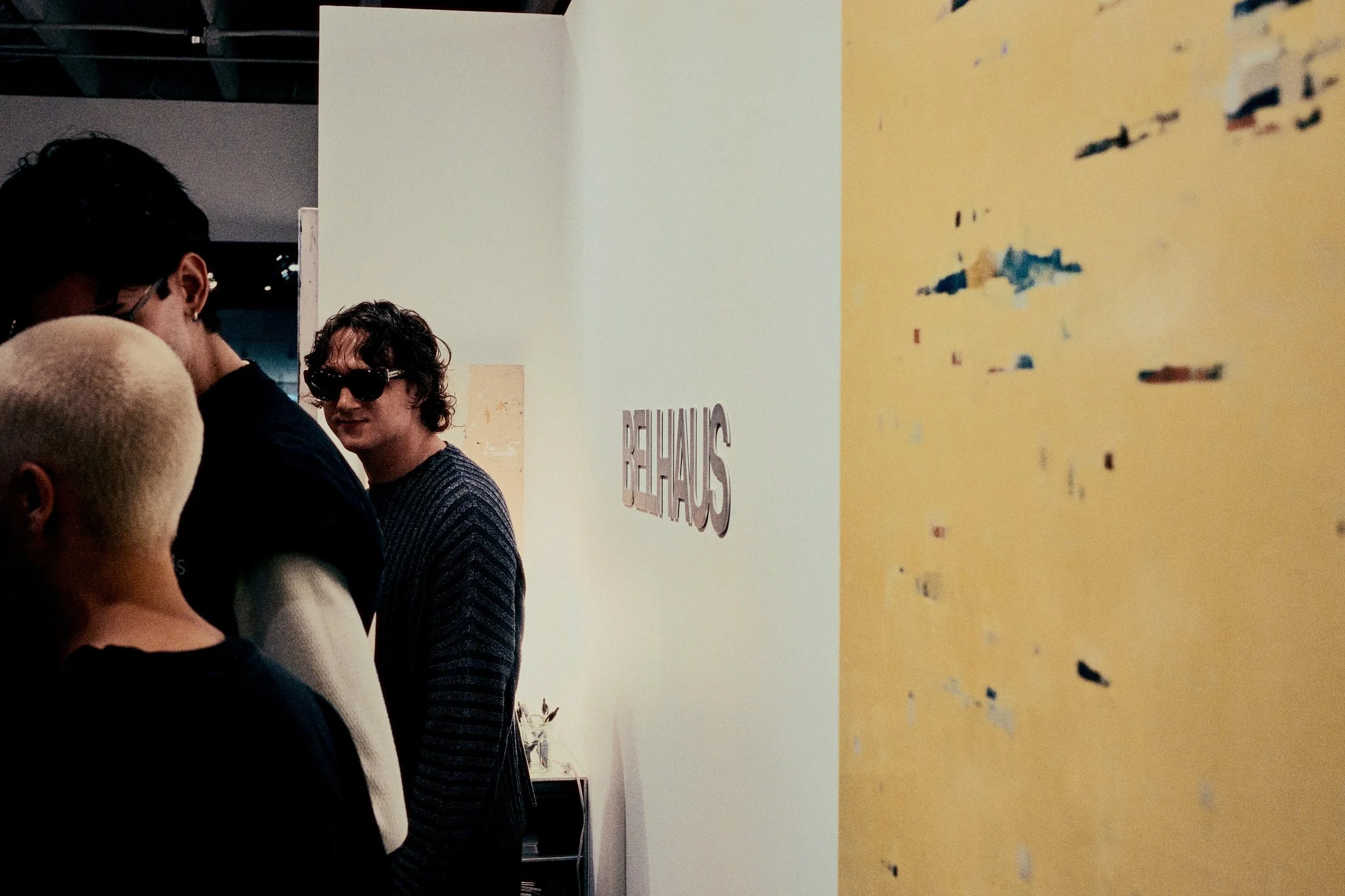 A group of people in an art gallery or exhibition space, with a sign that says 'BETHAUS' on a white wall in the background. One person is wearing sunglasses, and others are engaged in conversation or looking at art.