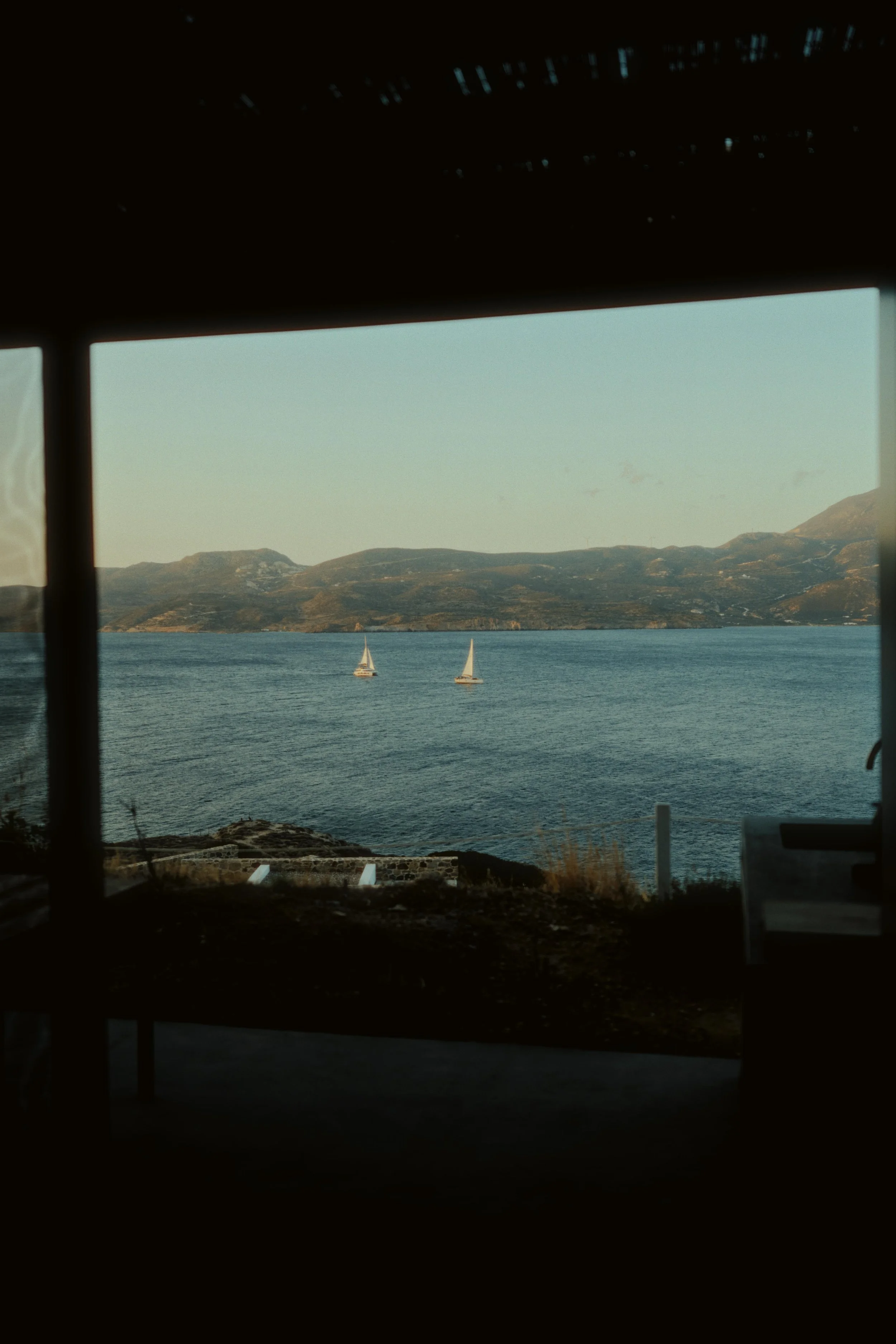 View of a lake with two sailboats sailing on calm water, framed by an open window or door, with mountains in the background.