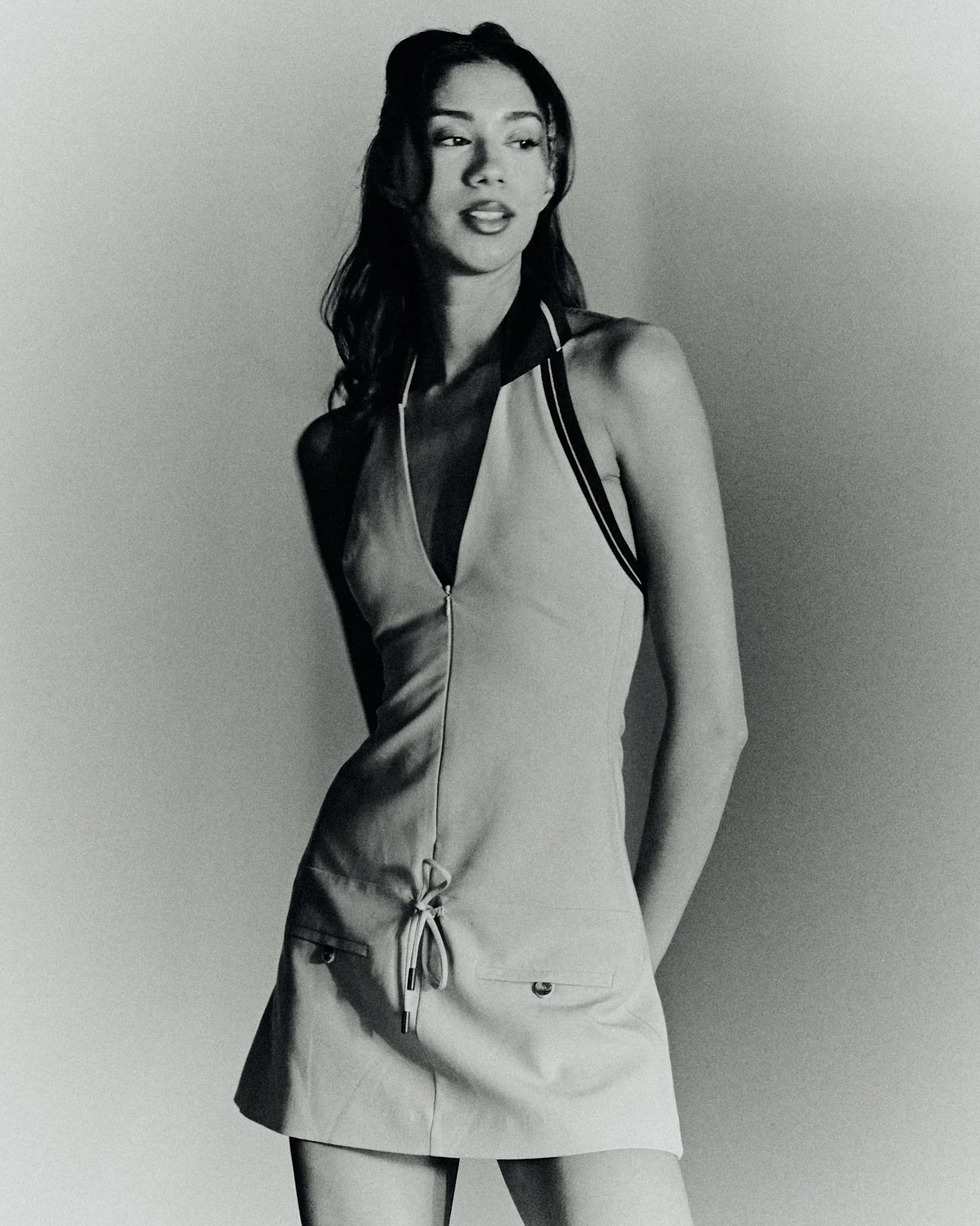 A woman in a sleeveless zip-up dress with a drawstring at the waist, standing against a plain background.