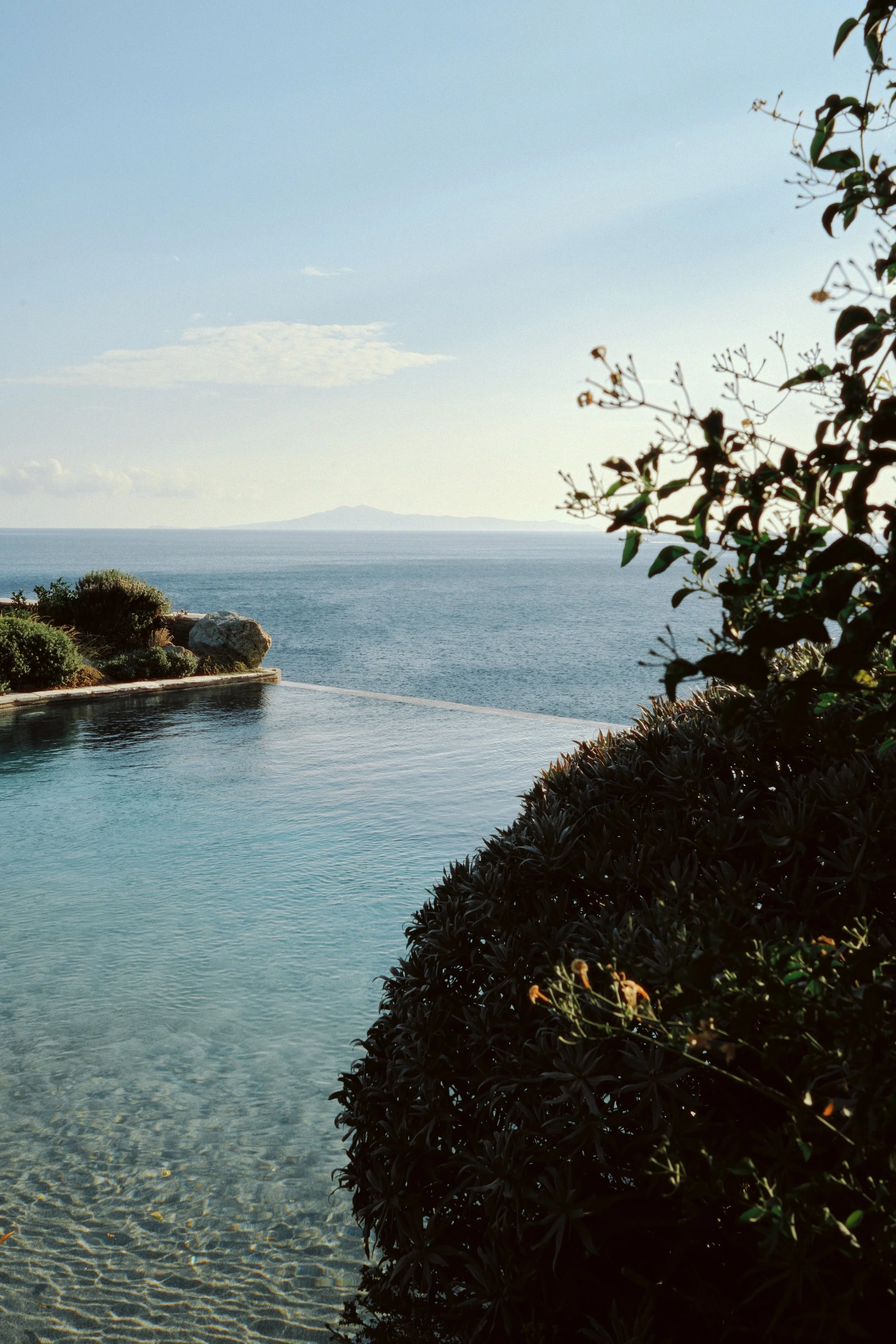 Ocean view with a pool in the foreground, shrubs and trees on the right side, and a distant island on the horizon under a partly cloudy sky.