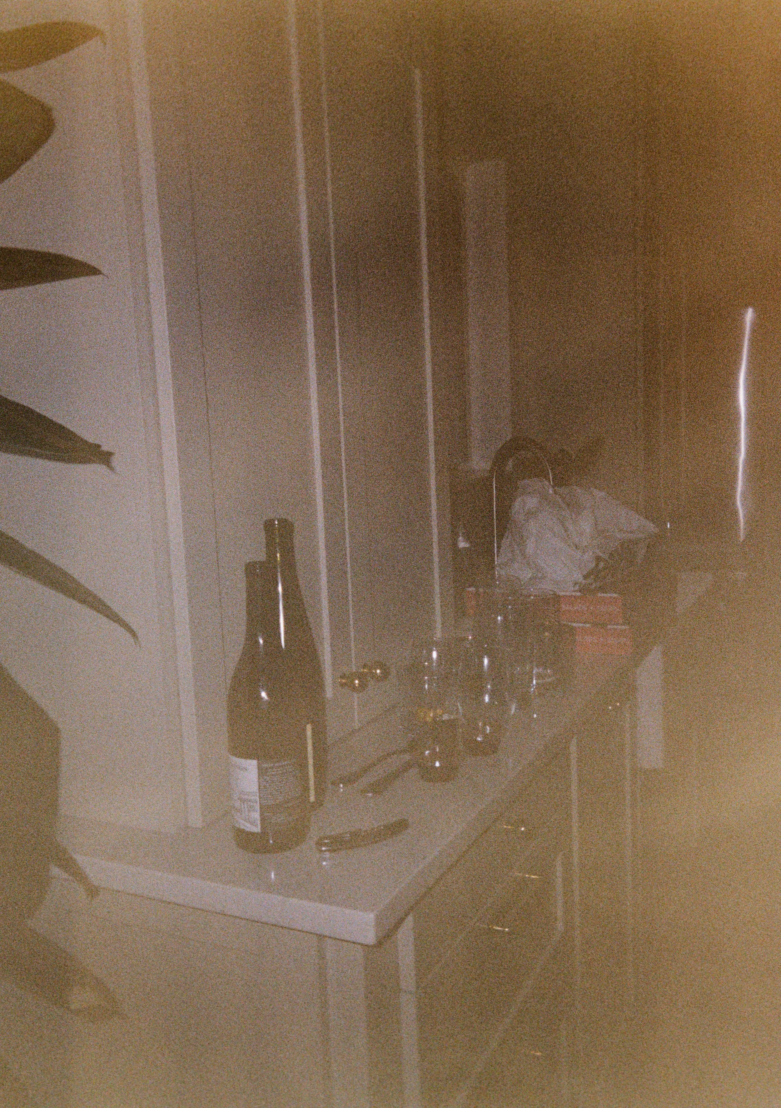 A kitchen counter with a beer bottle, glasses, and a utility knife, with a person in the background wearing headphones and a gray hoodie.