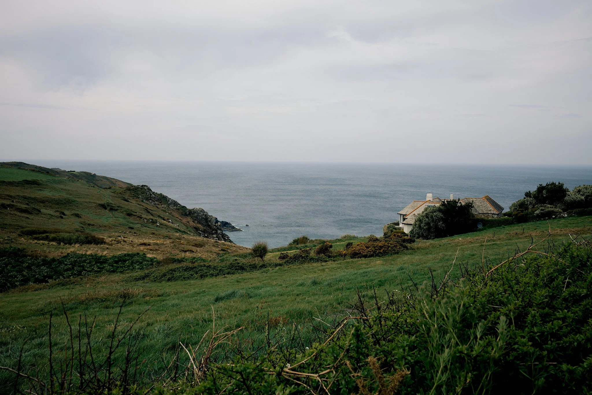 A coastal landscape with green grassy hills, a house, and the ocean in the background under a cloudy sky.