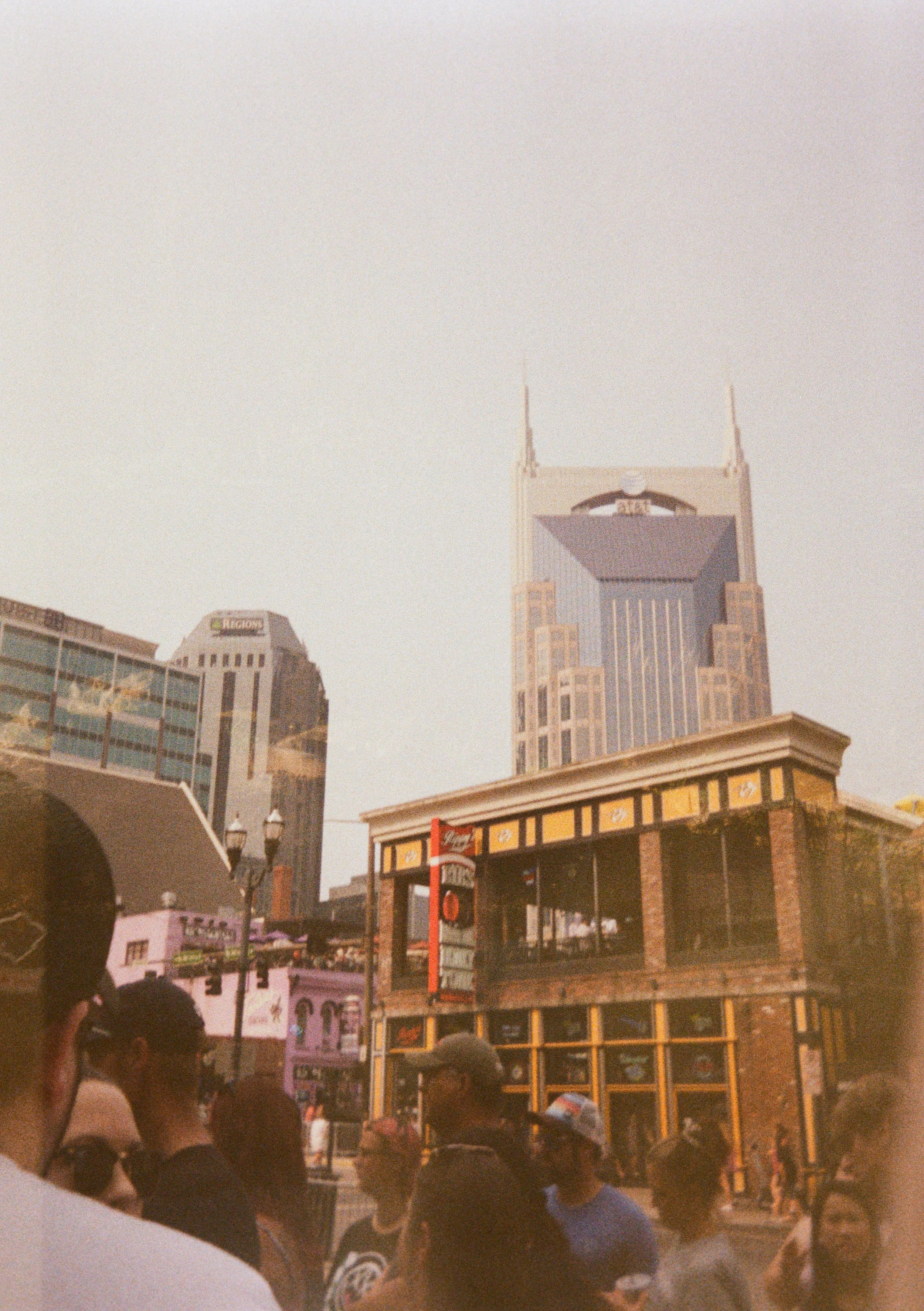 Crowd of people in urban street scene with tall buildings and city skyline in the background, including a recognizable twin-spired building.