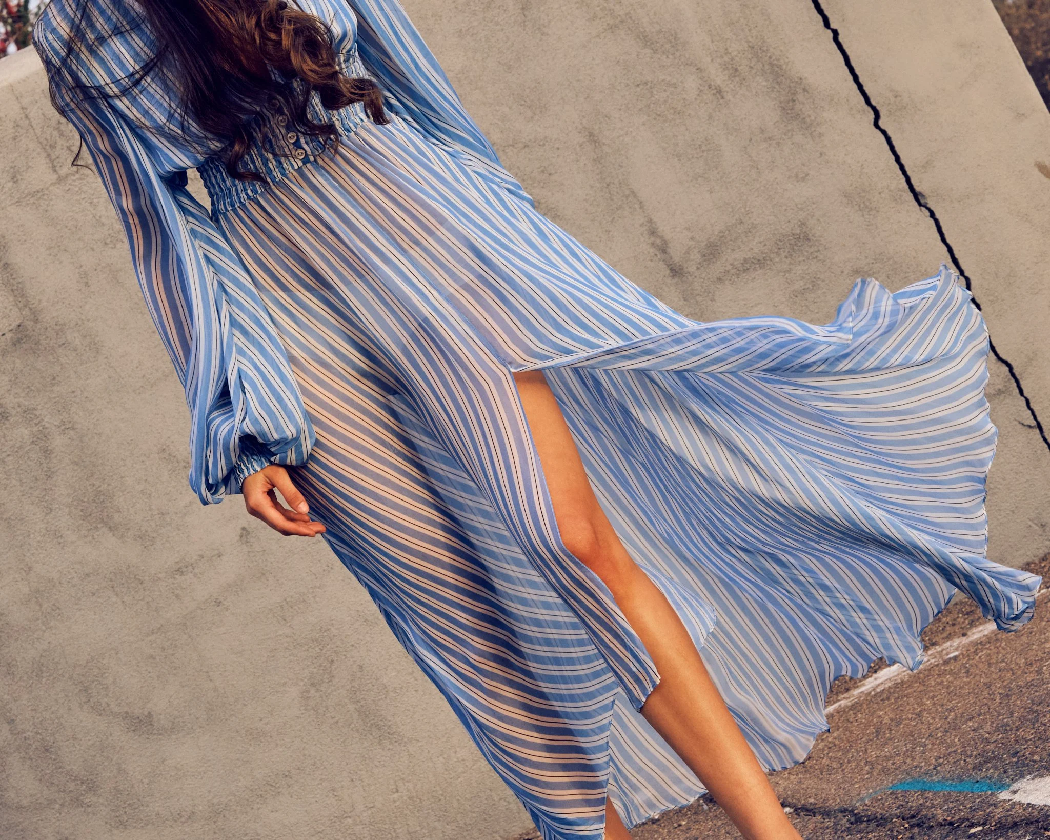 A woman wearing a flowing, blue and white striped dress with a slit, standing against a textured concrete wall.