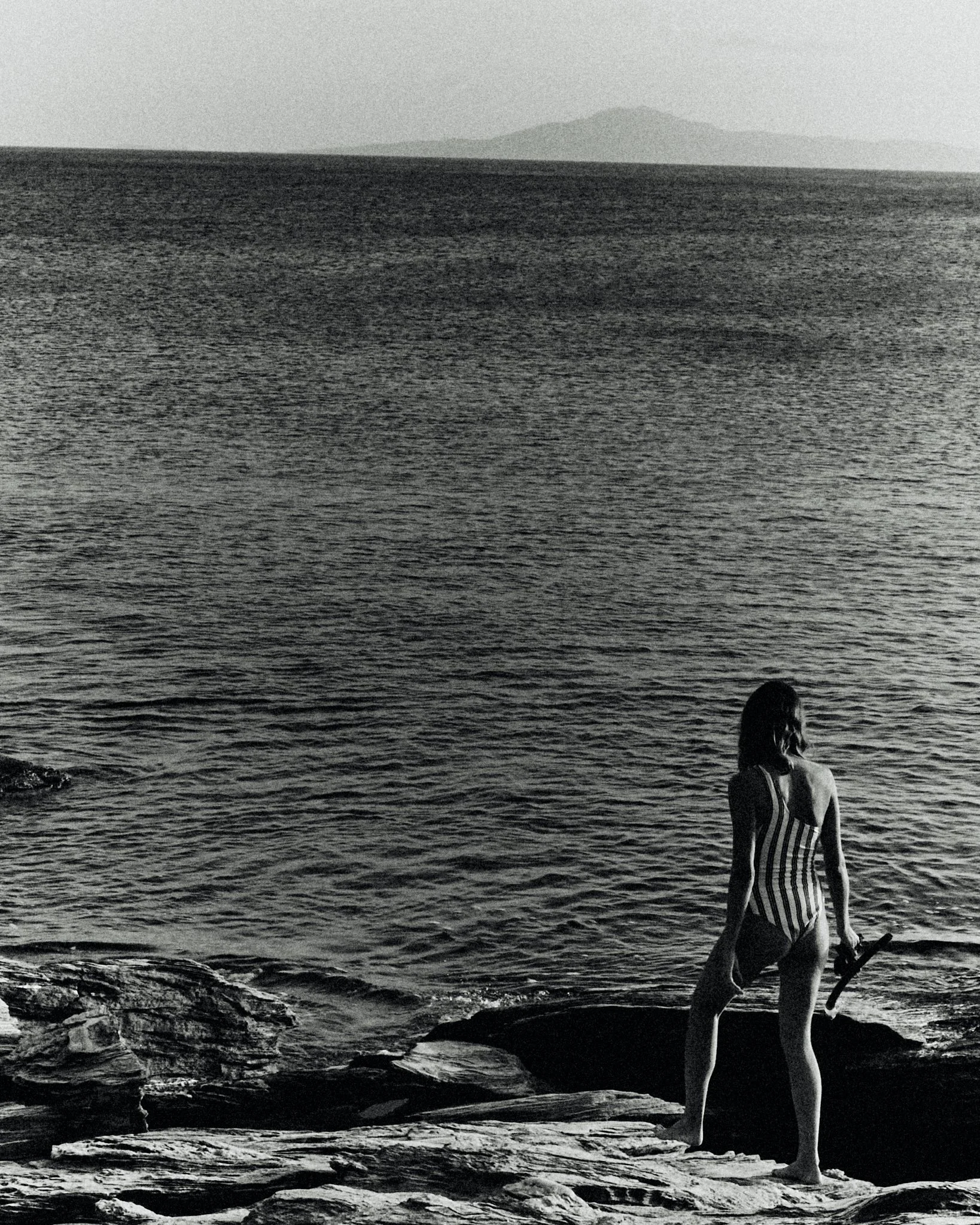 A woman standing on rocks by the water, holding a shoe, with a large body of water and distant land in the background, in black and white.