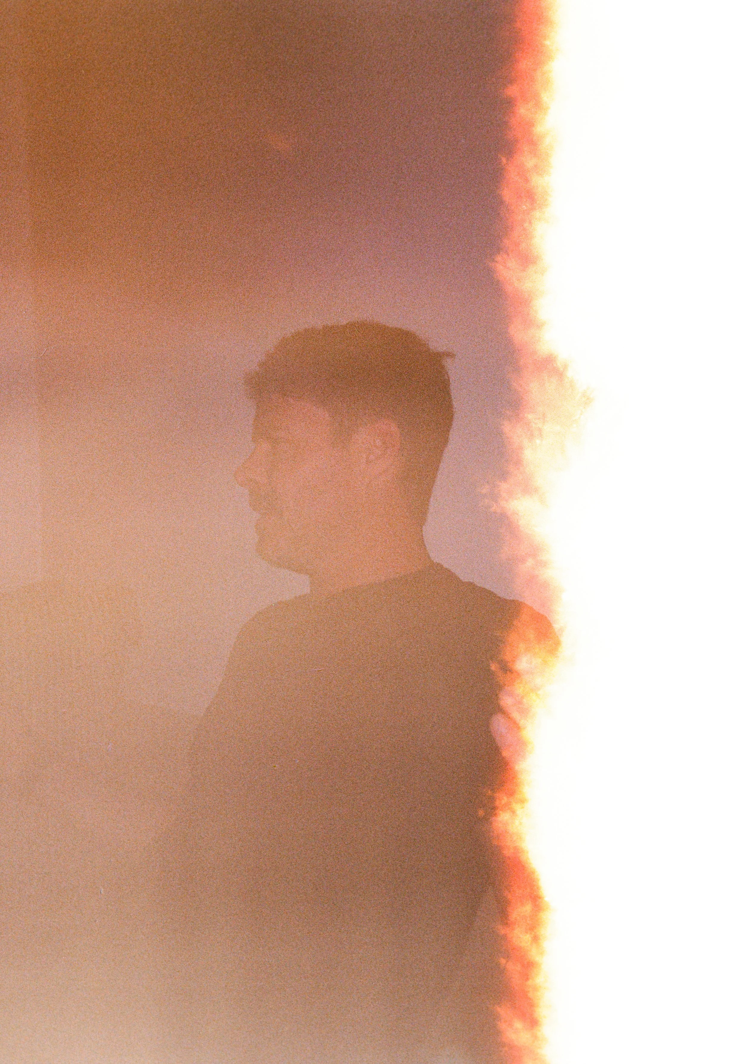 Profile of a young man with short hair, wearing a dark shirt, against a colorful, blurred background with a light flare on the right side.