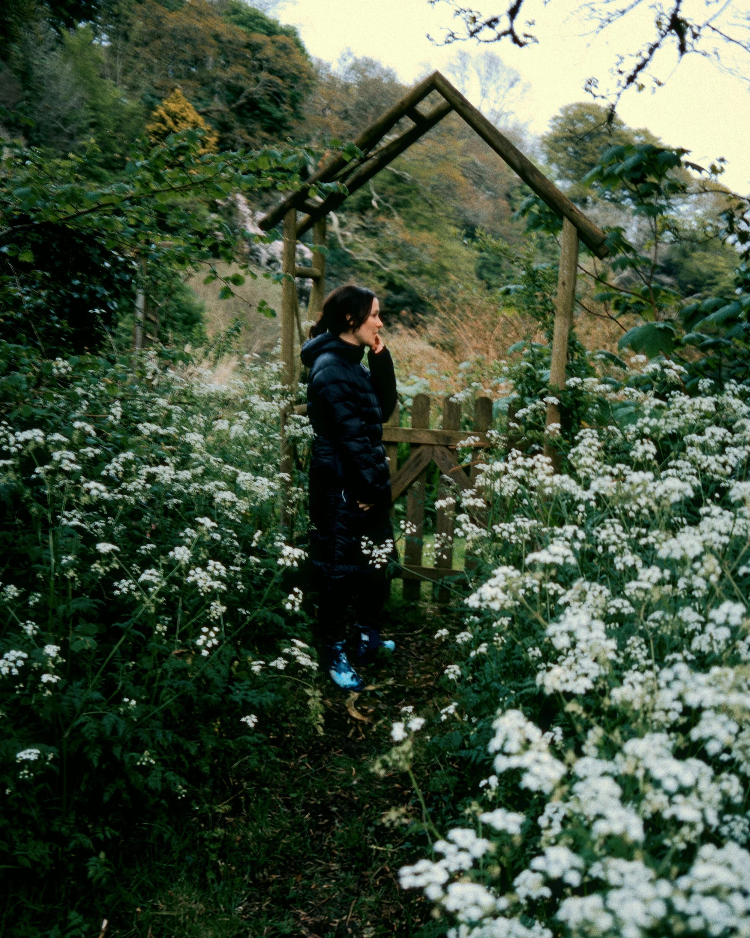 Woman in black puffy coat standing on a narrow garden path surrounded by white flowers, near a small wooden gate and archway, with trees and hills in the background.