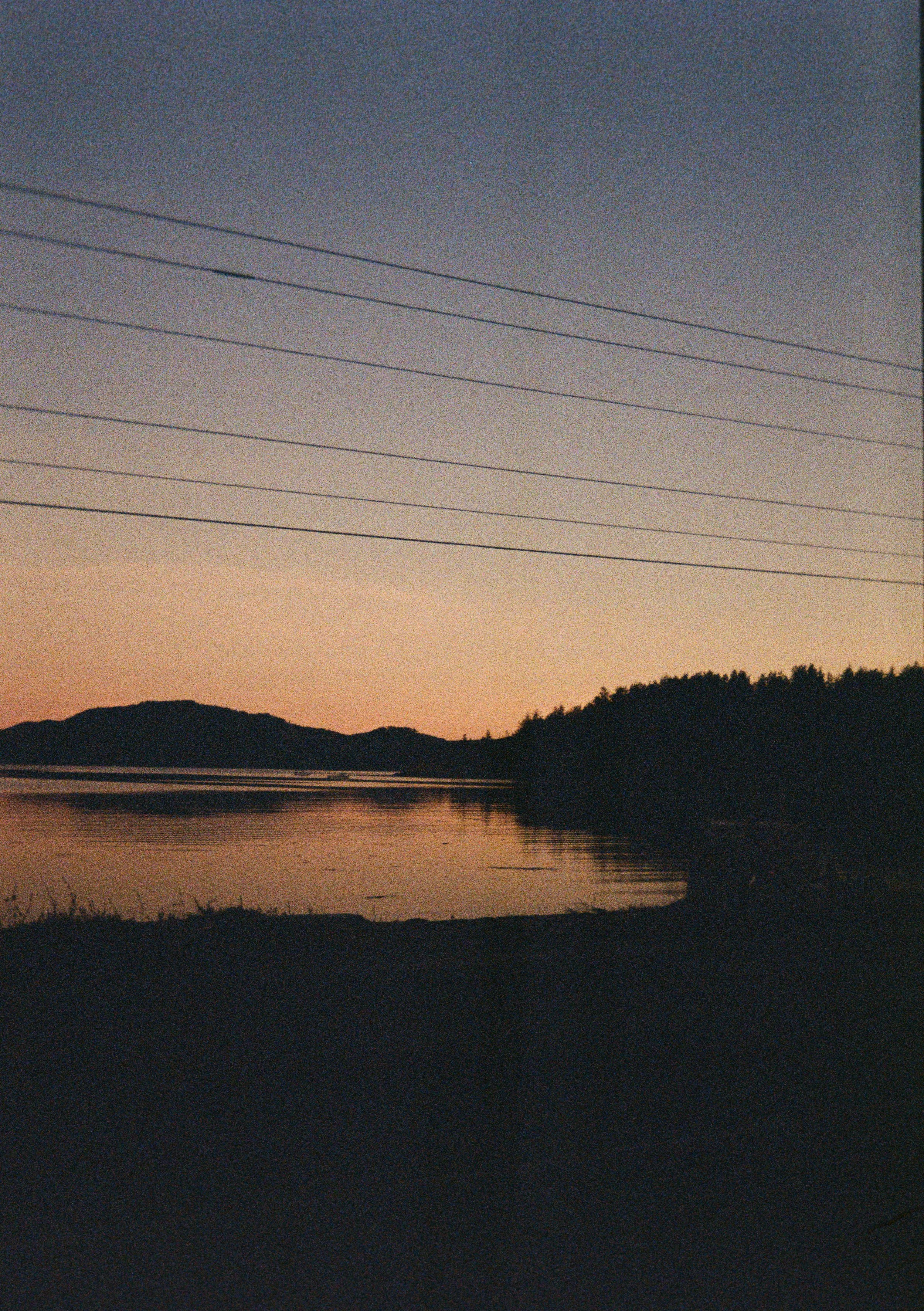 A serene landscape featuring a calm body of water with hills in the background, under a twilight sky with power lines overhead.