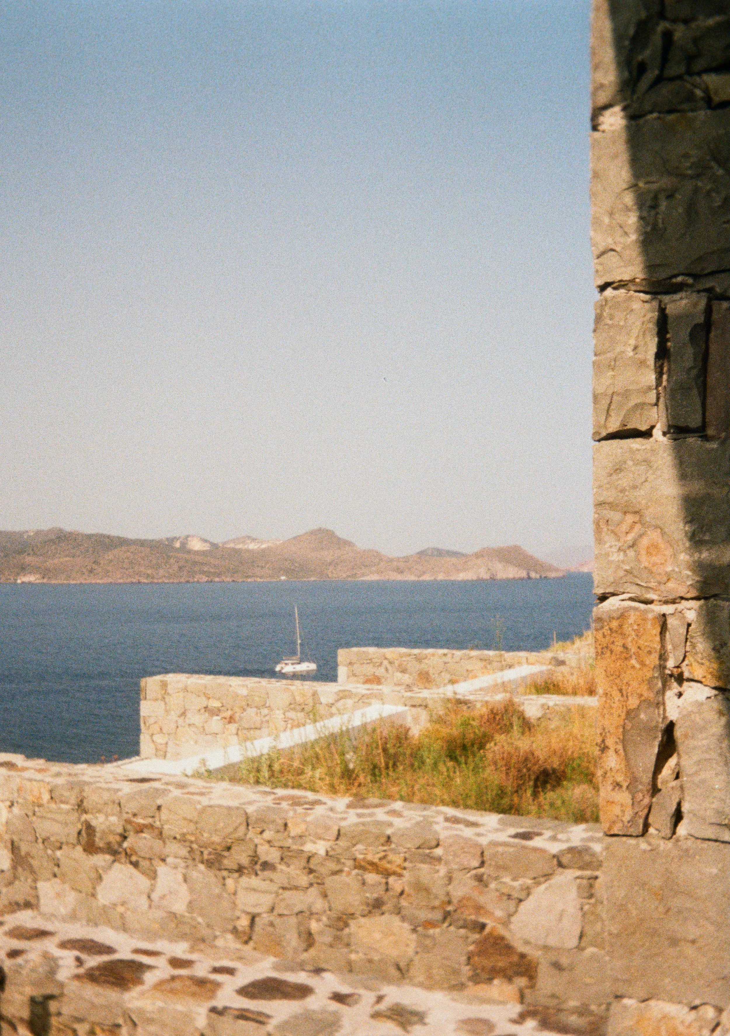 Scenic view of a body of water with a small sailboat, stone walls, and distant mountains, seen through a stone window.
