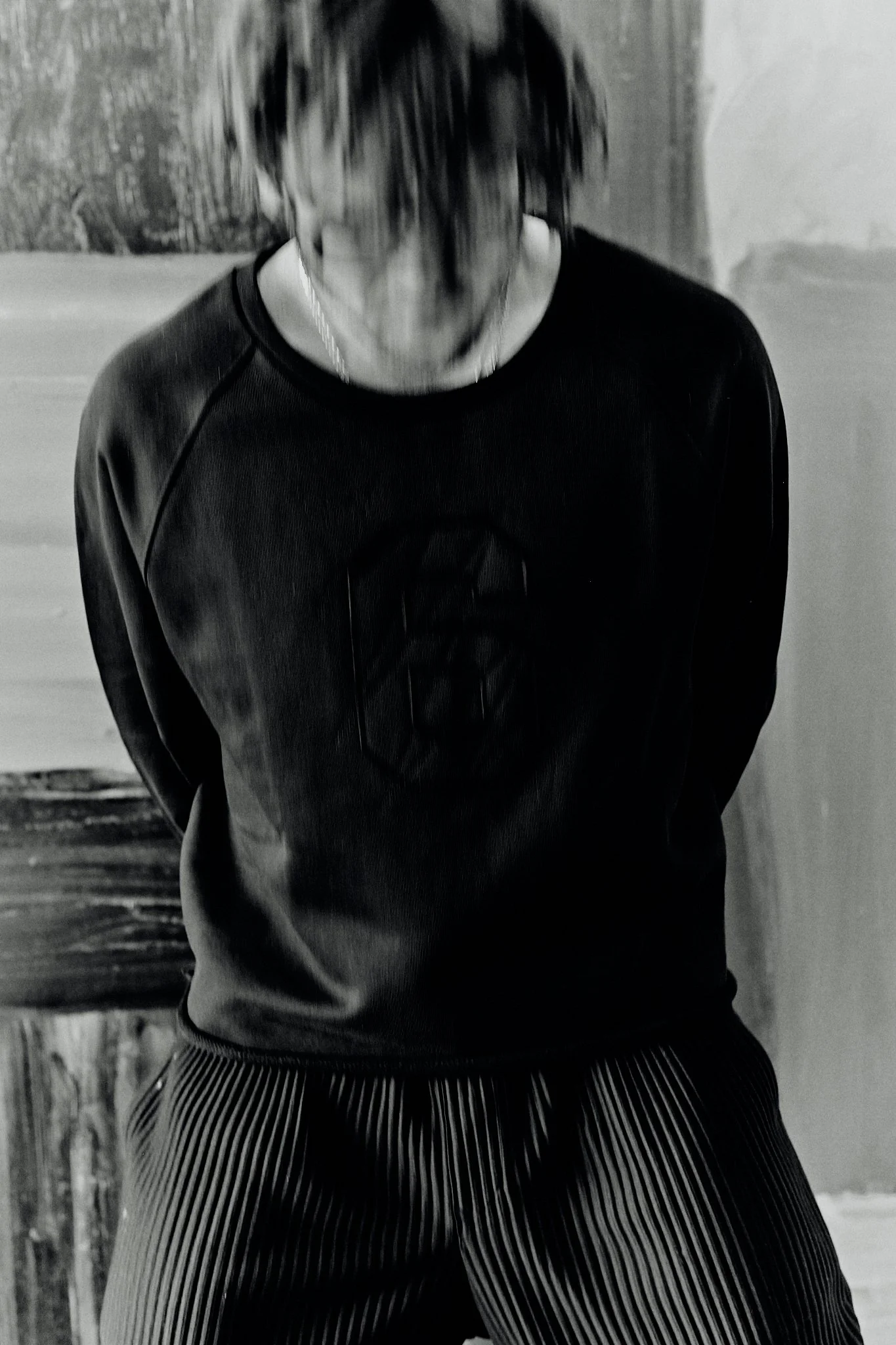 A person with curly hair wearing a dark sweatshirt with a logo and striped pants, standing indoors against a wall.
