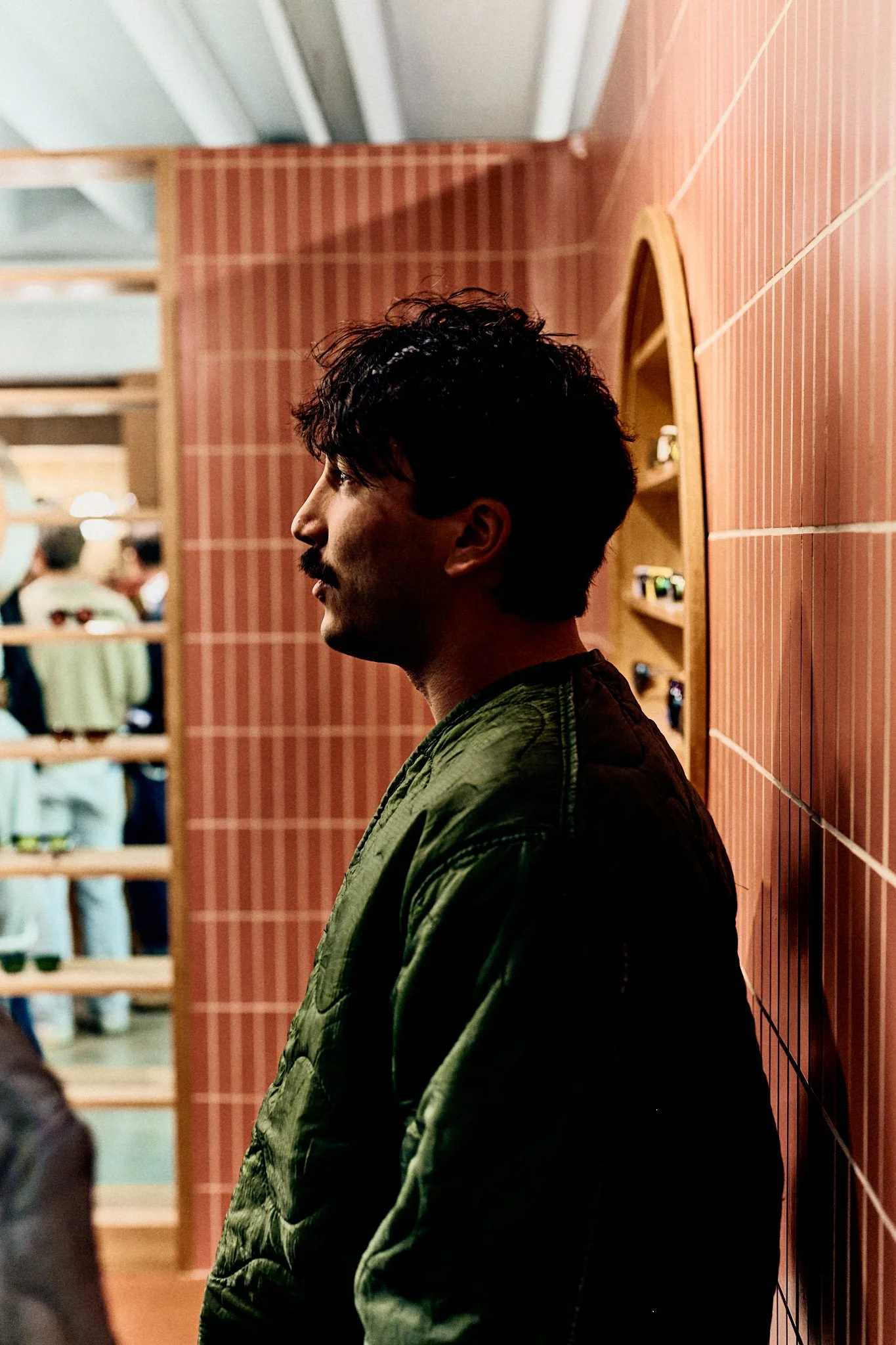 A man with dark, curly hair and a mustache, wearing a green quilted jacket, leaning against a red-tiled wall in an indoor setting, with people in the background.