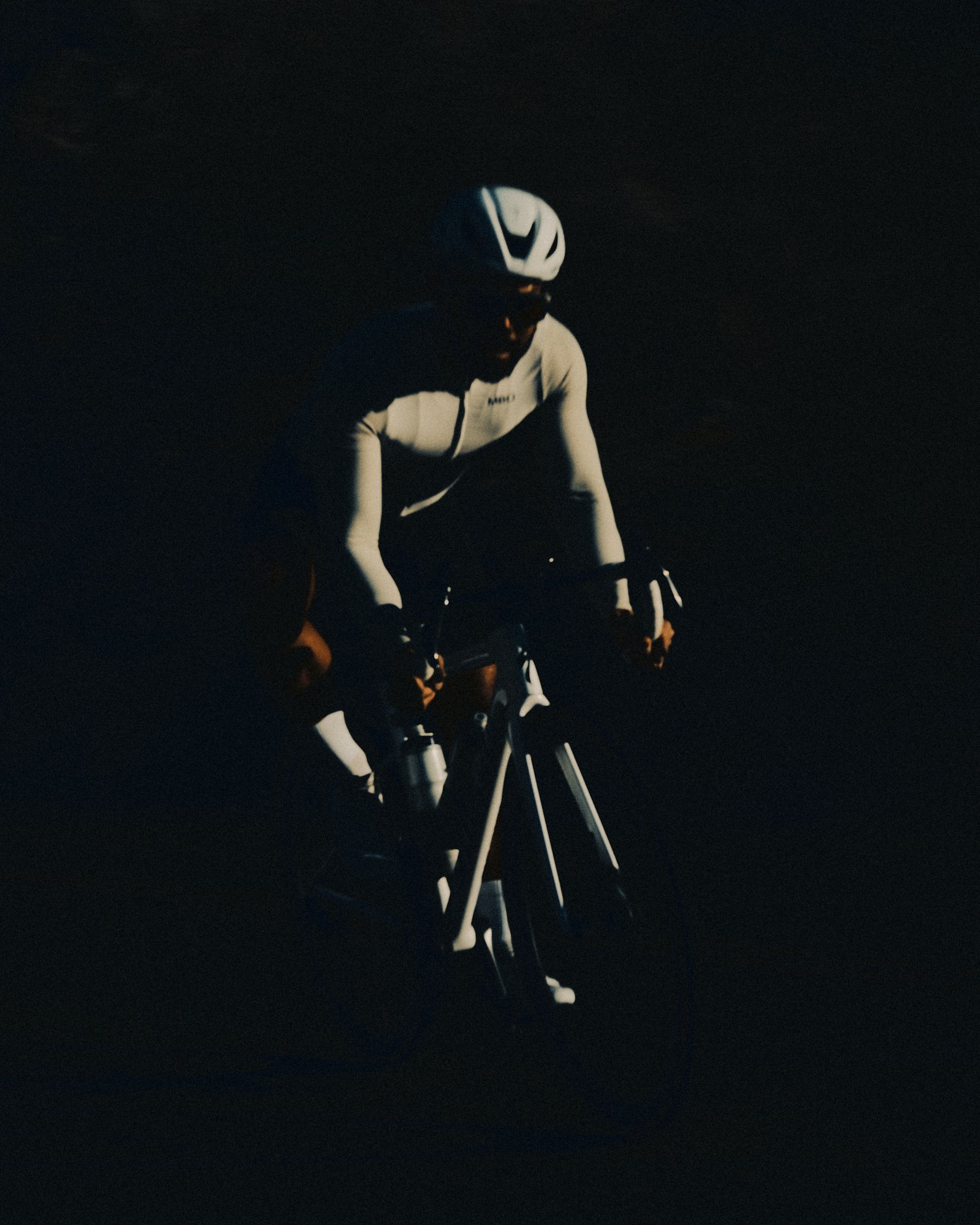 A cyclist wearing a white helmet and white long-sleeve shirt riding a bicycle in darkness.