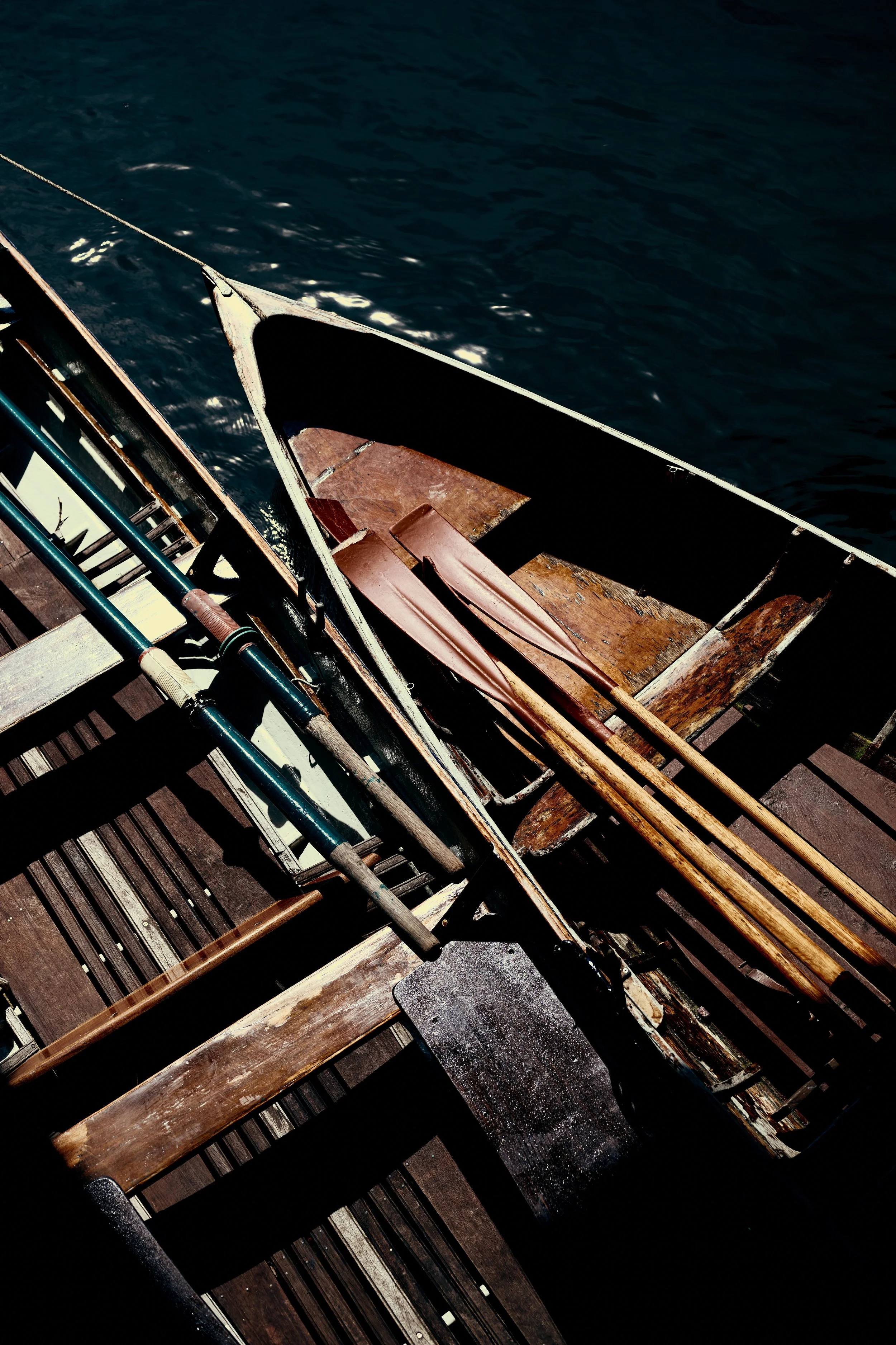 Two wooden oars resting on a wooden dock next to a small rowboat with an outboard motor, viewed from above over dark blue water.
