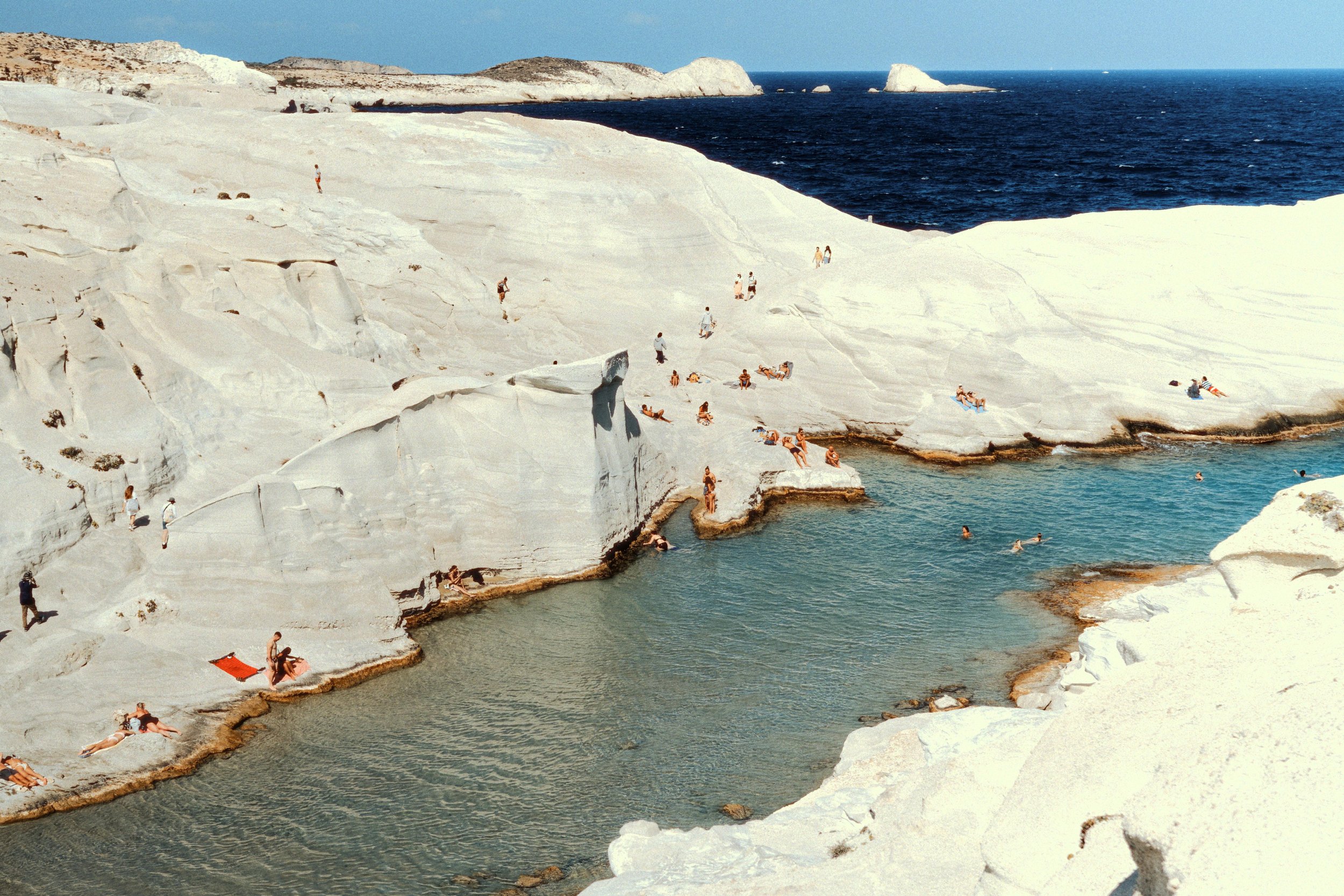 People sunbathing and swimming in a small natural pool surrounded by white rock formations on a sunny day, with the ocean in the background.