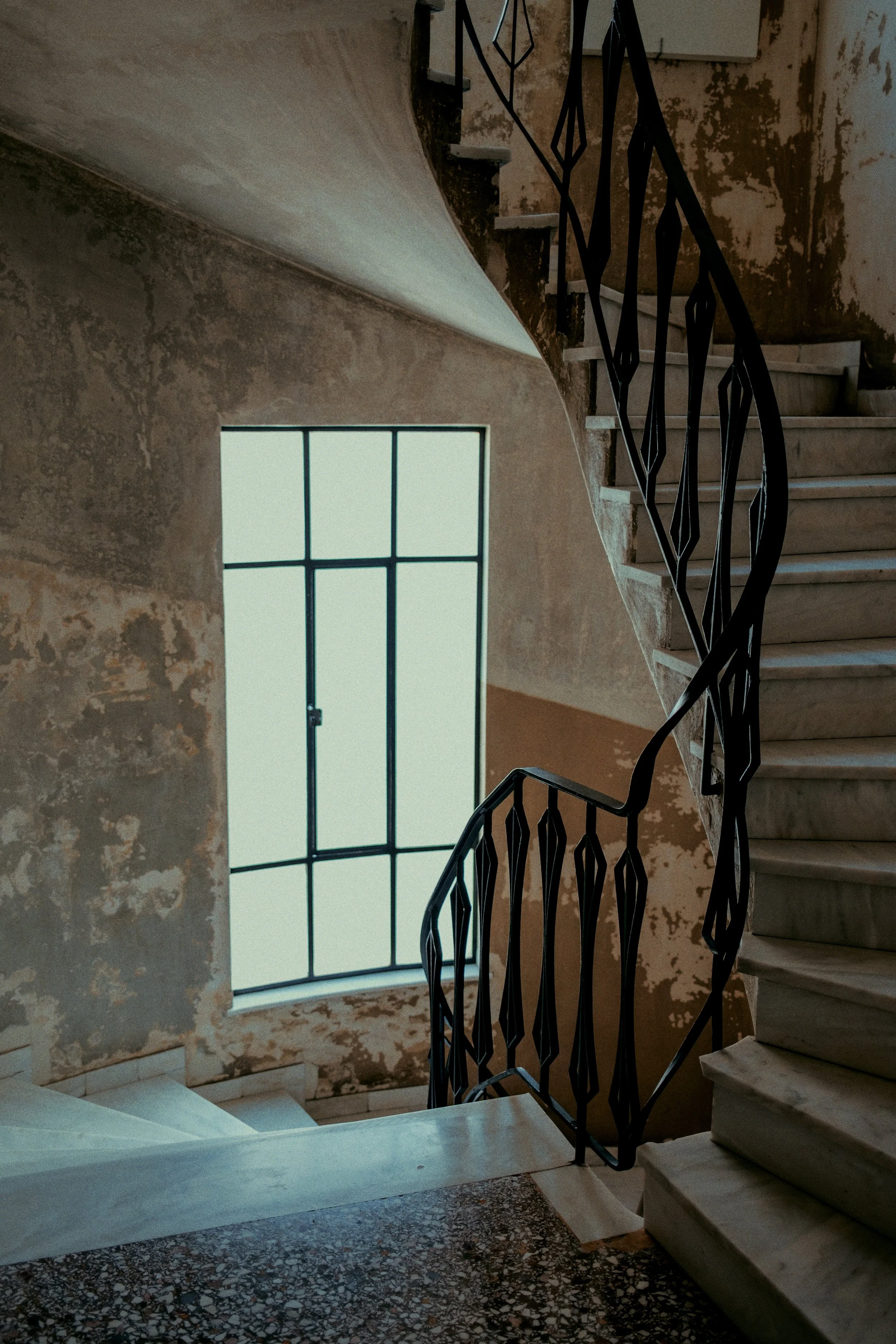 Interior of an old building with a marble staircase, metal railing, and a large window with a grid pattern, showing peeling paint and weathered walls.
