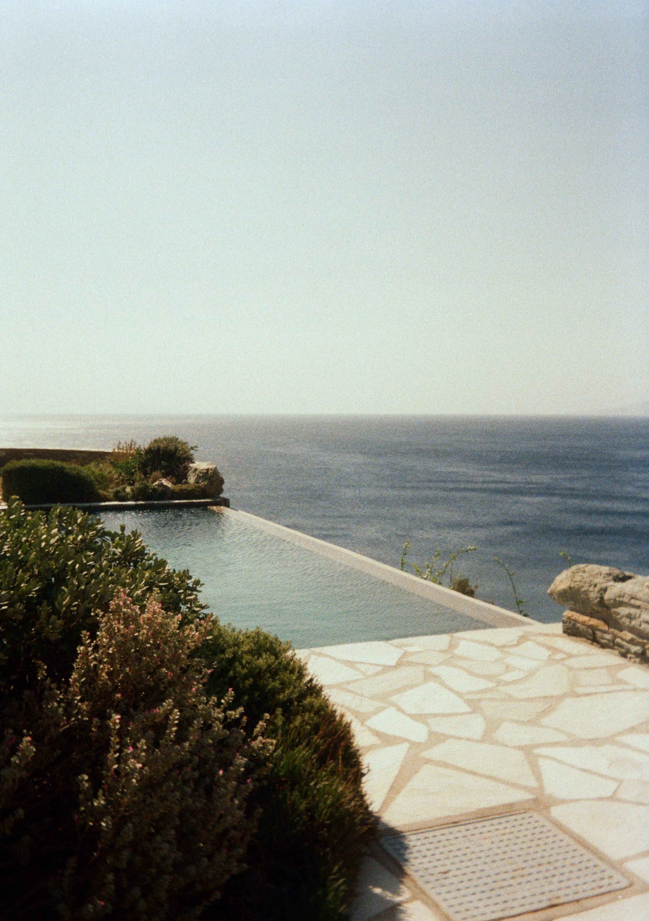 View of an infinity pool overlooking the ocean with plants and rocks nearby on a sunny day.
