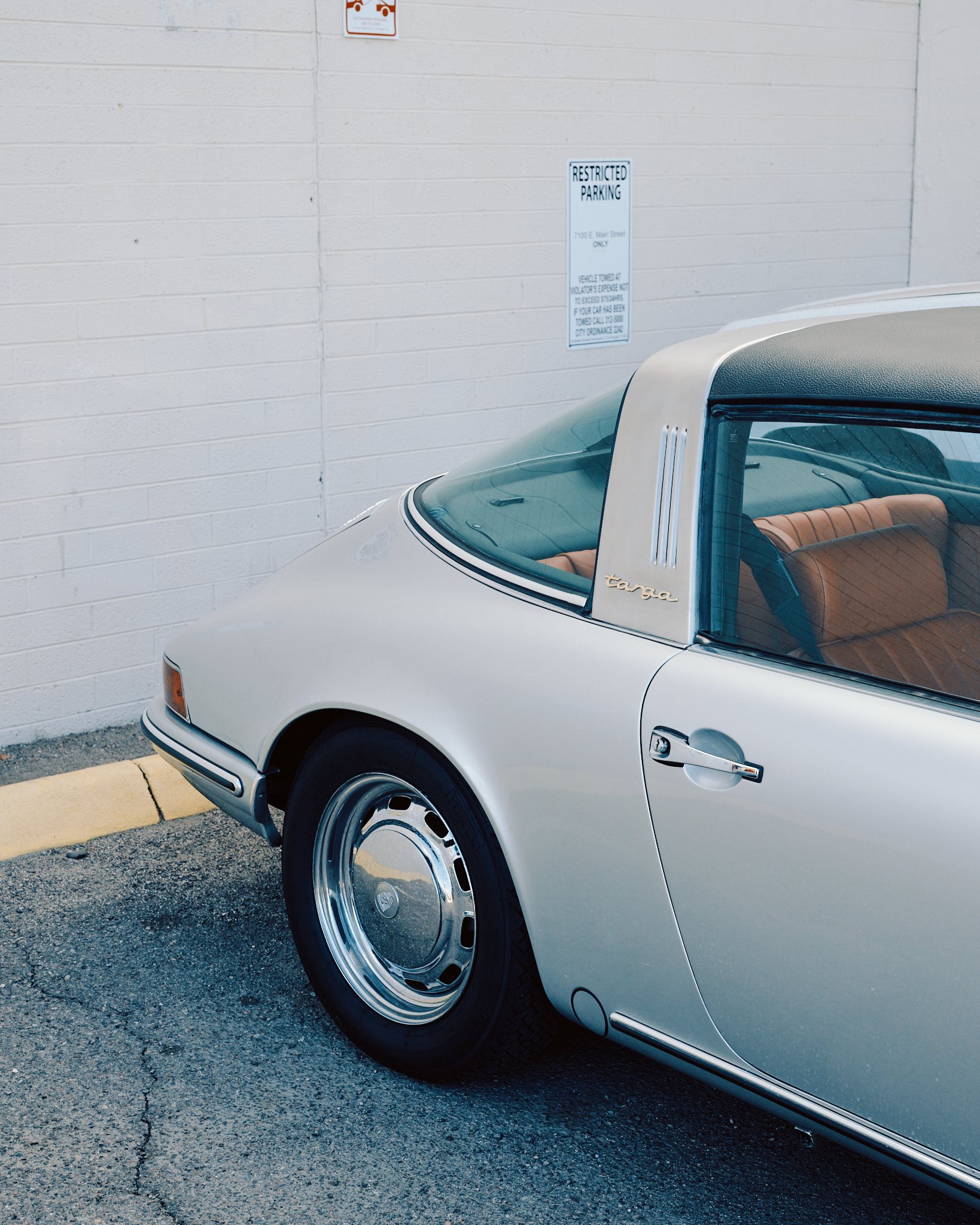 Rear side of a vintage silver Porsche Targa parked near a white brick wall with a restricted parking sign.
