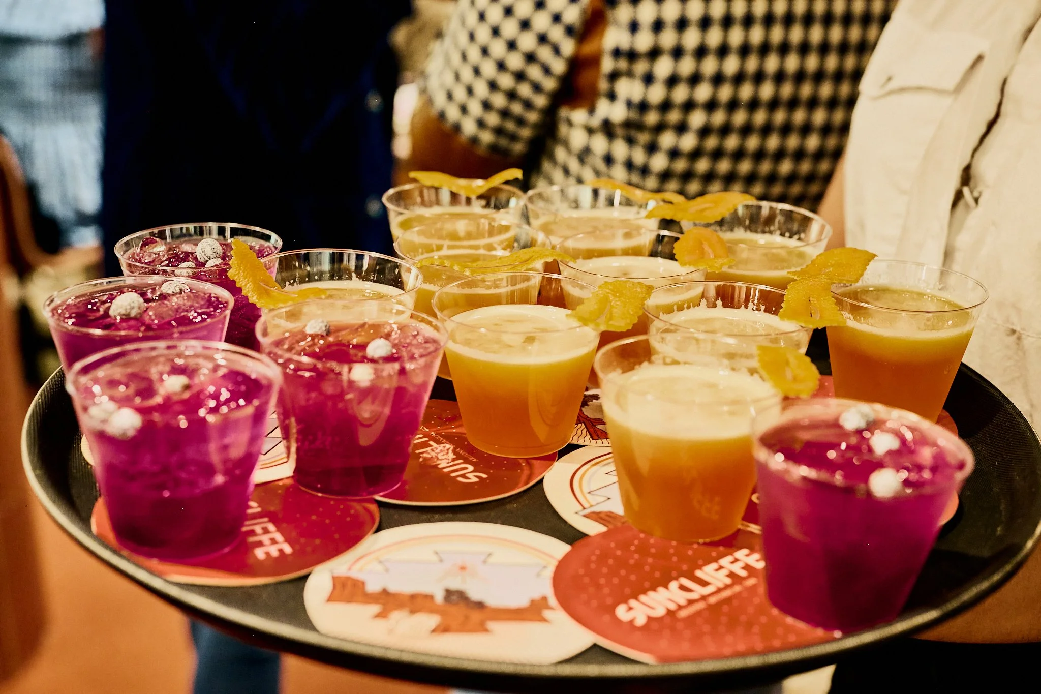 A tray of colorful drinks, including purple and yellow beverages, garnished with lemon twists, on a table at a social gathering.