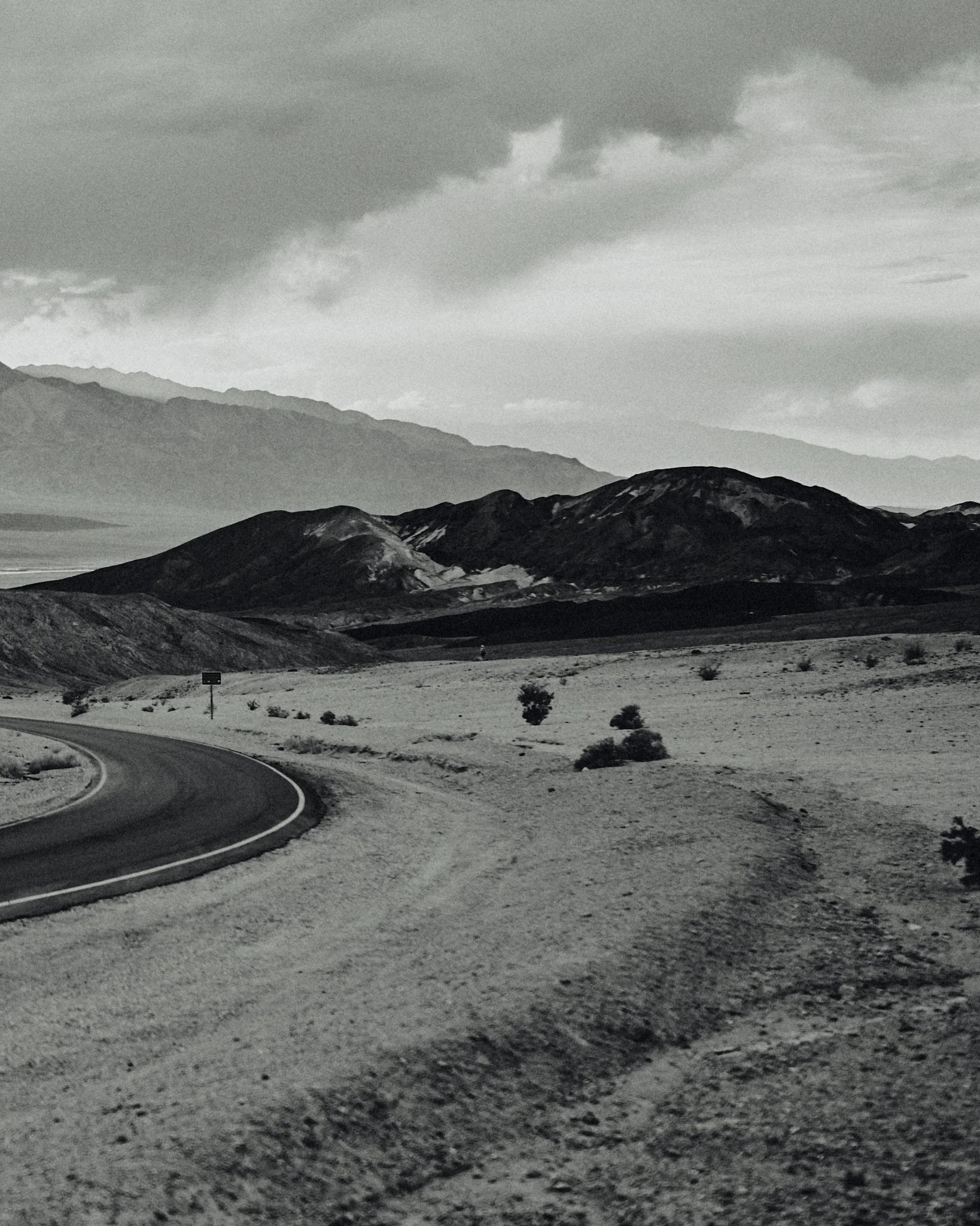 A black and white photograph of a winding desert road with mountains in the background and cloudy sky overhead.