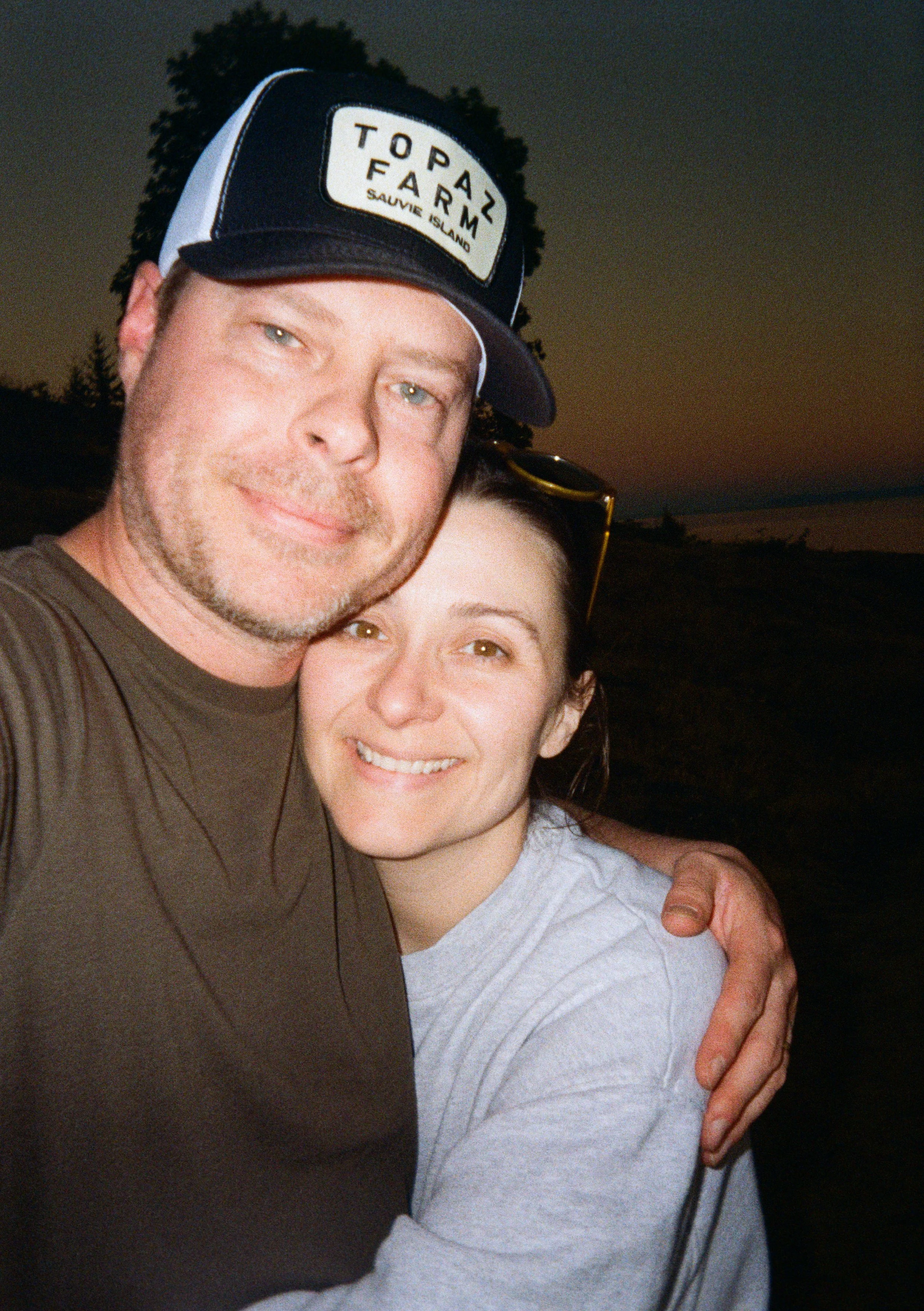A man and woman smiling and hugging outdoors at sunset, the man wearing a dark baseball cap and the woman wearing sunglasses on her head.