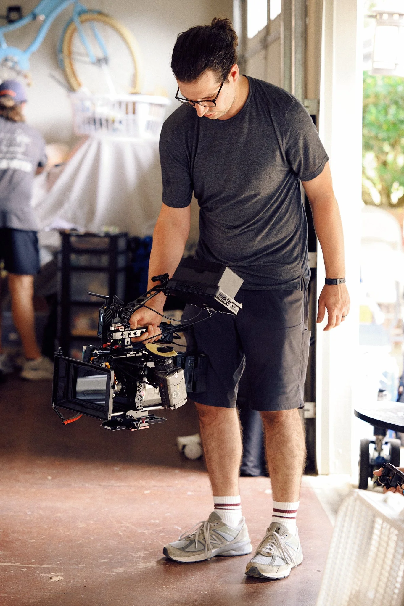 A man wearing glasses, a dark gray t-shirt, black shorts, white sneakers, and striped socks is holding a professional video camera, standing inside a bright, casual space. There is a person in the background working at a table and a white bicycle mou