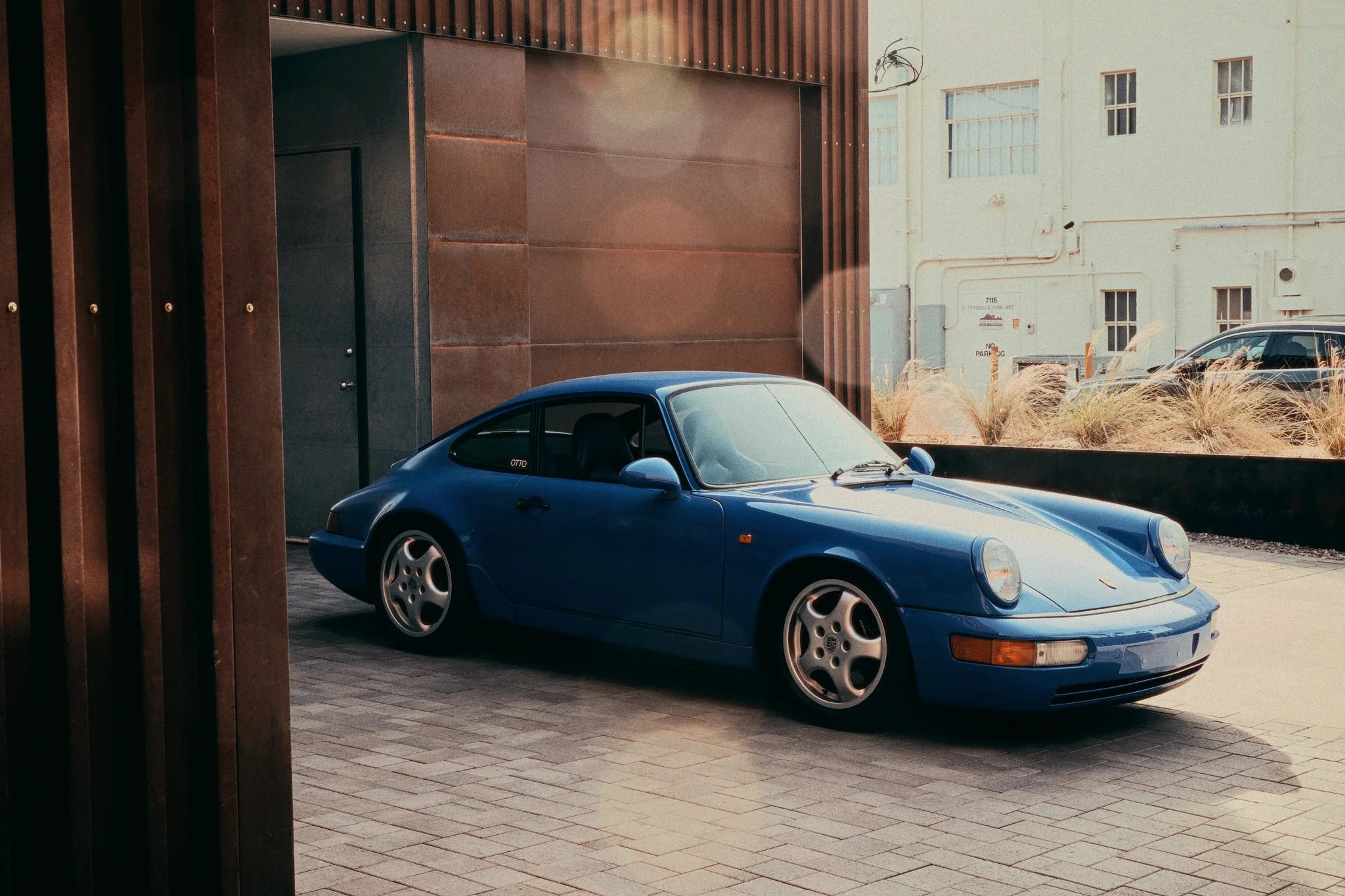 A blue vintage Porsche 911 parked outside a building with modern architecture and tall windows.