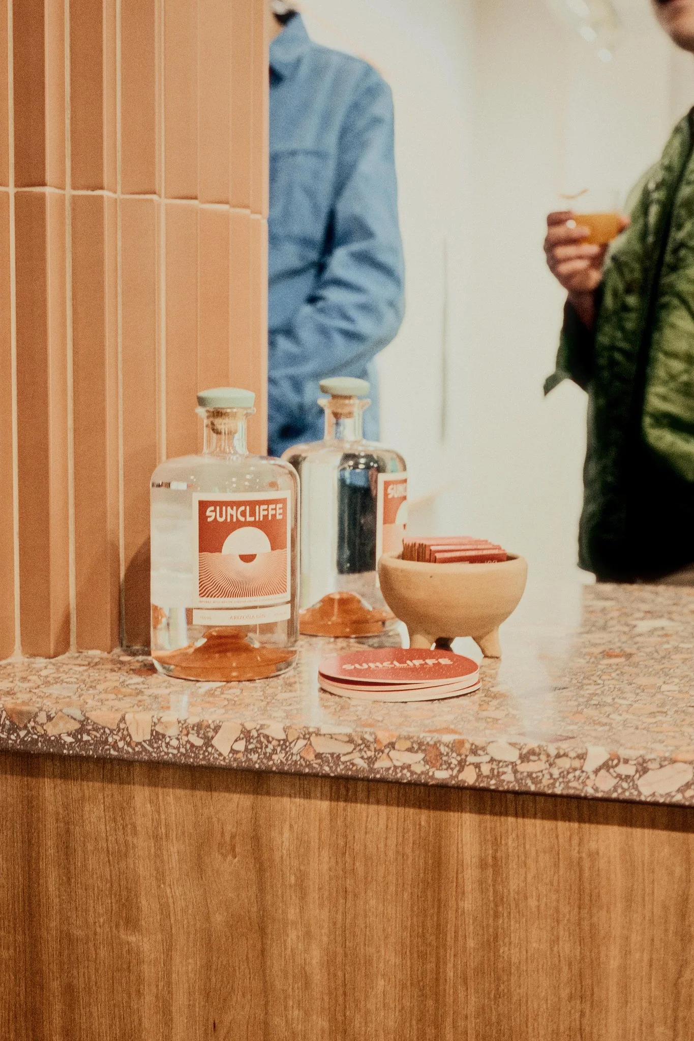 Coffee shop counter with Suncliffe branded bottles, napkins, and people holding cups.