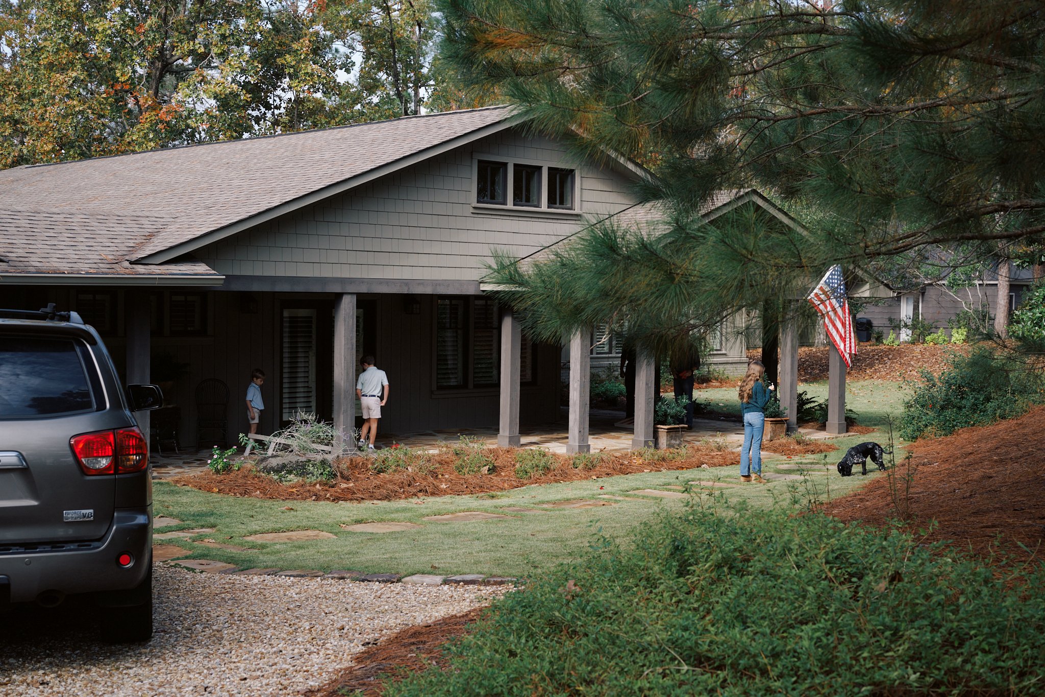 People standing on the porch of a house with a front yard, an American flag, and a black dog on a leash in the yard. A large vehicle is parked in the driveway. The house is surrounded by trees and landscaping.