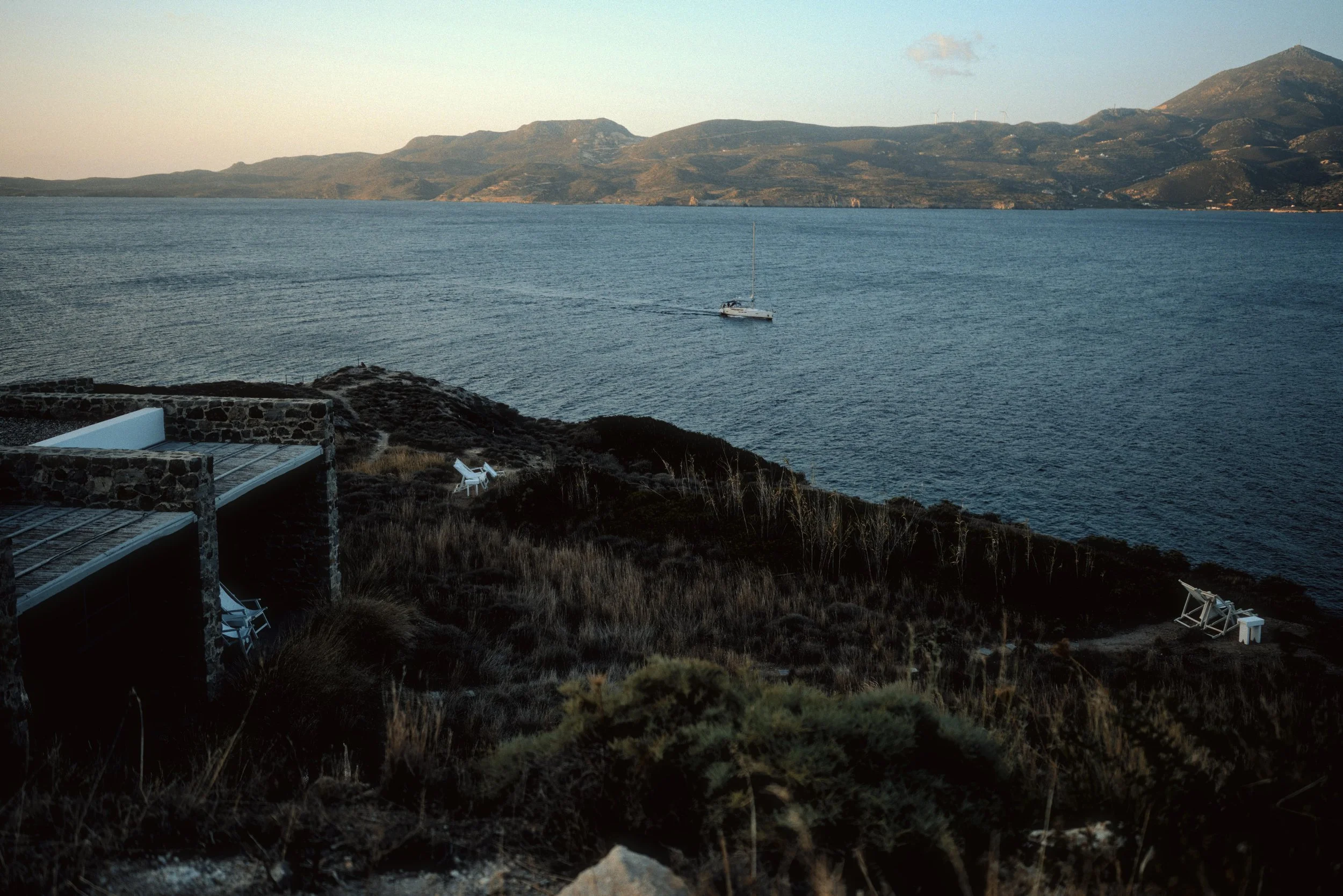 A tranquil coastal scene with a sailboat on calm water, a rocky shoreline with a building, and hills in the background, during sunset or late afternoon.