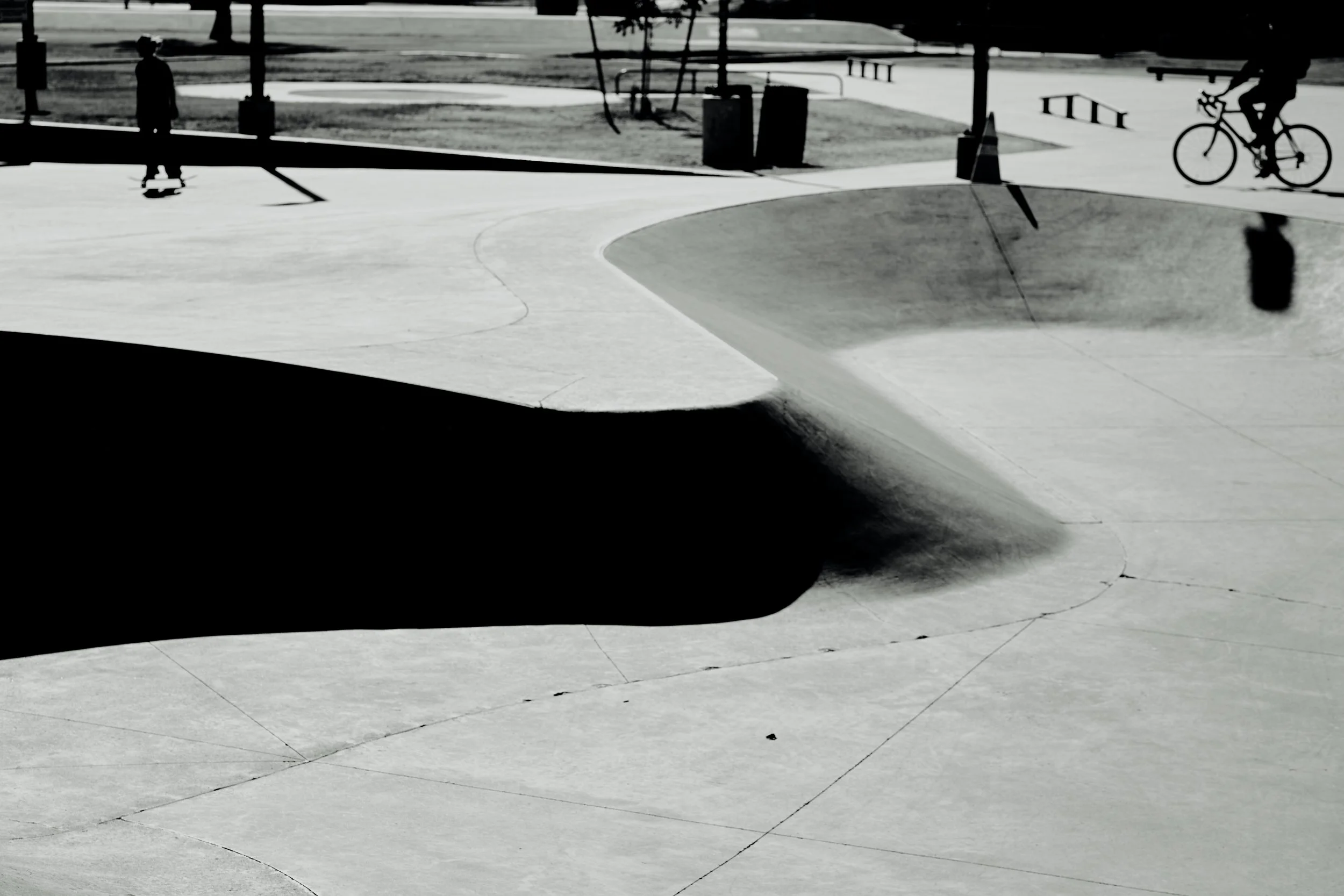 Black and white photo of a skatepark with a large bowl, a skateboarder, and a cyclist in the background, with trees and picnic tables in the distance.