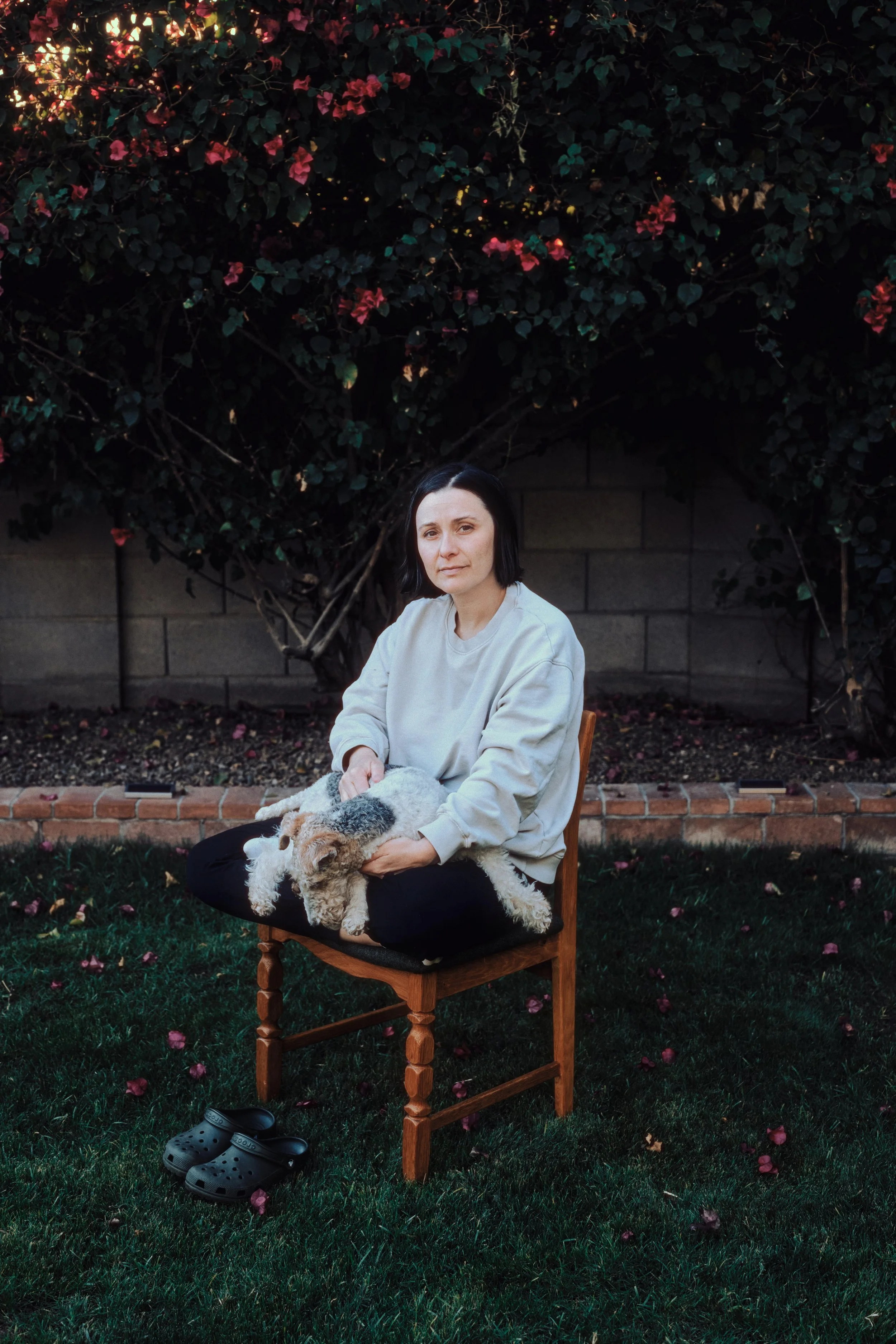 A woman with short dark hair sitting on a wooden chair outdoors, holding a small white and brown dog in her lap, with black Crocs on the grass nearby, and a large bush with pink flowers in the background.
