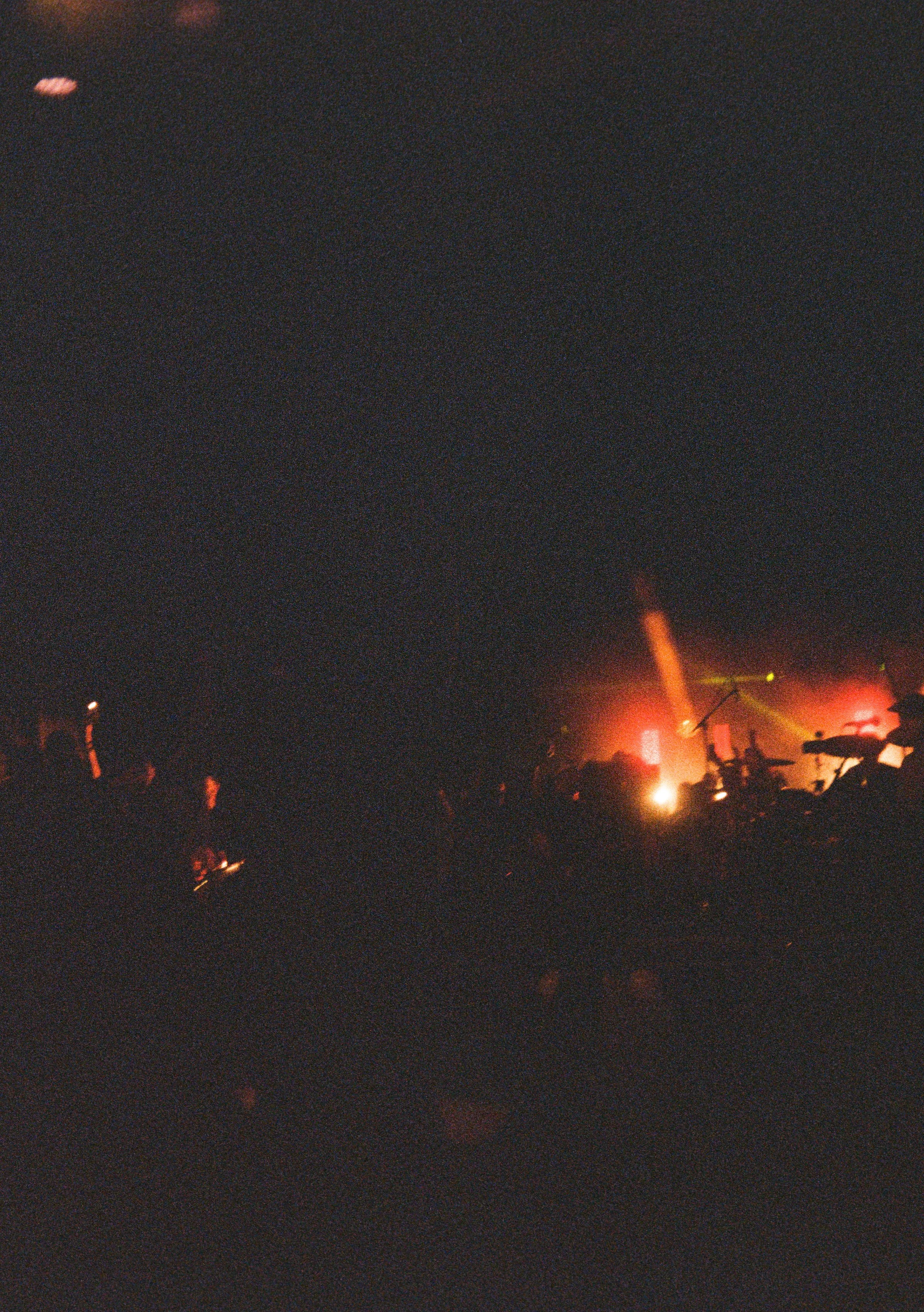Nighttime scene with silhouettes of people gathered around a fire, motorcycles, and fire trucks in the background, with emergency lights illuminating the scene.