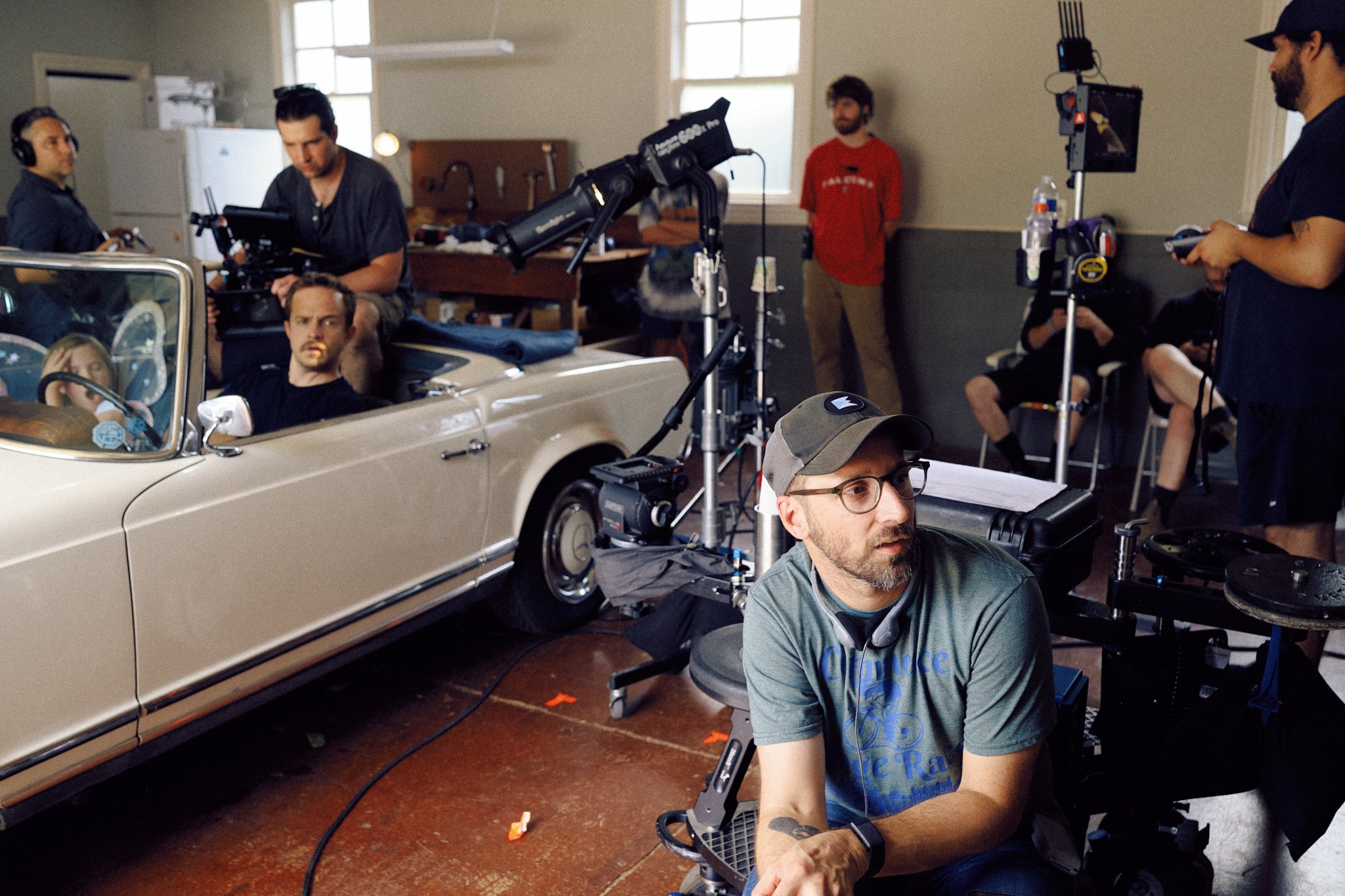 Behind-the-scenes of a film set with a vintage beige convertible car, crew members operating cameras and equipment, and actors sitting inside the car.