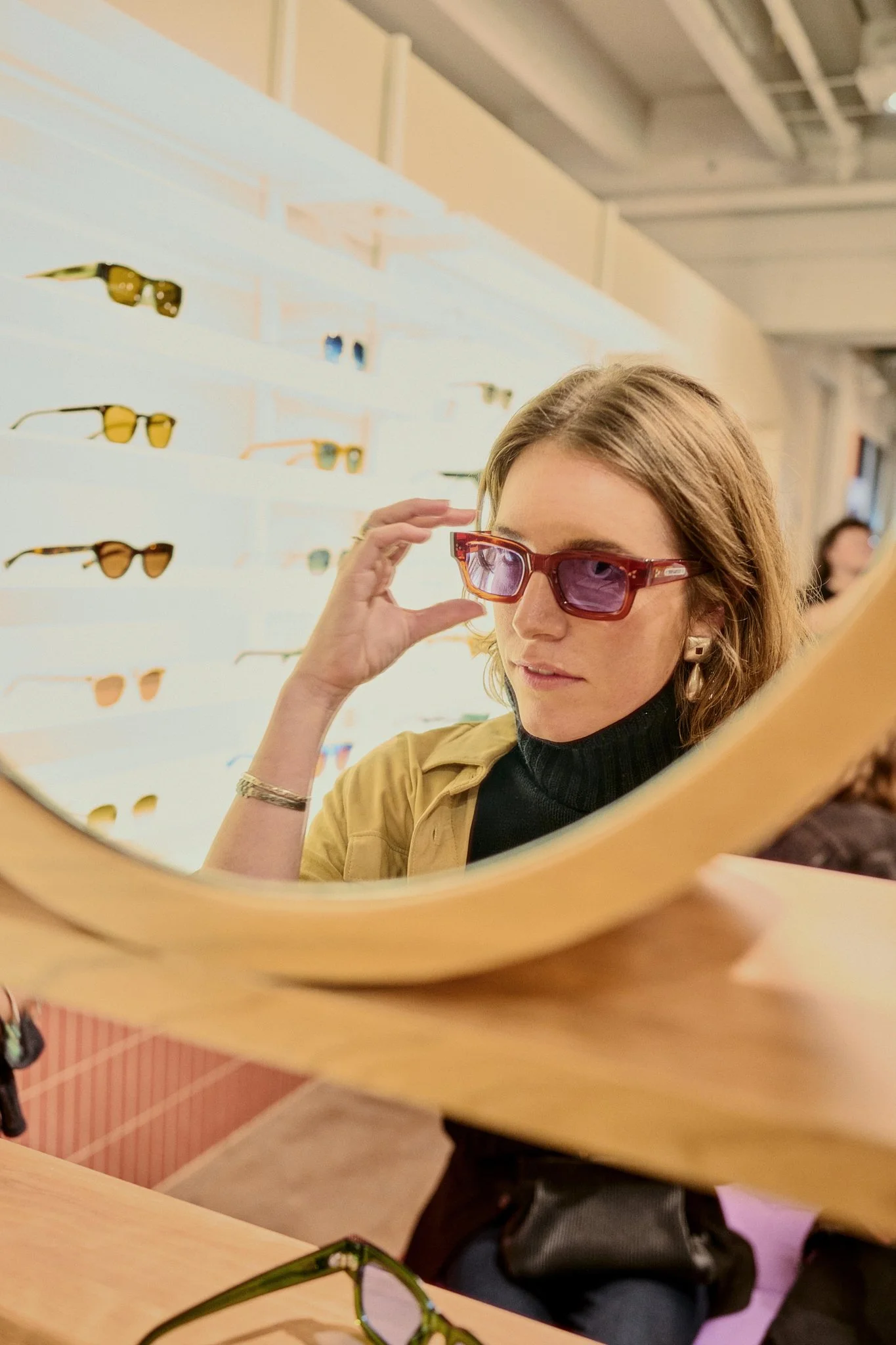 A woman trying on sunglasses in a store, seen through a mirror. She is wearing a black turtleneck, tan jacket, and statements earrings, with a wall of sunglasses displayed behind her.