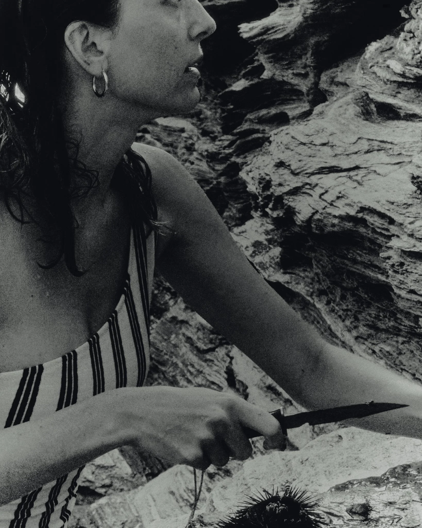 A woman wearing a striped top and earrings holding a knife, sitting near rocks and water.