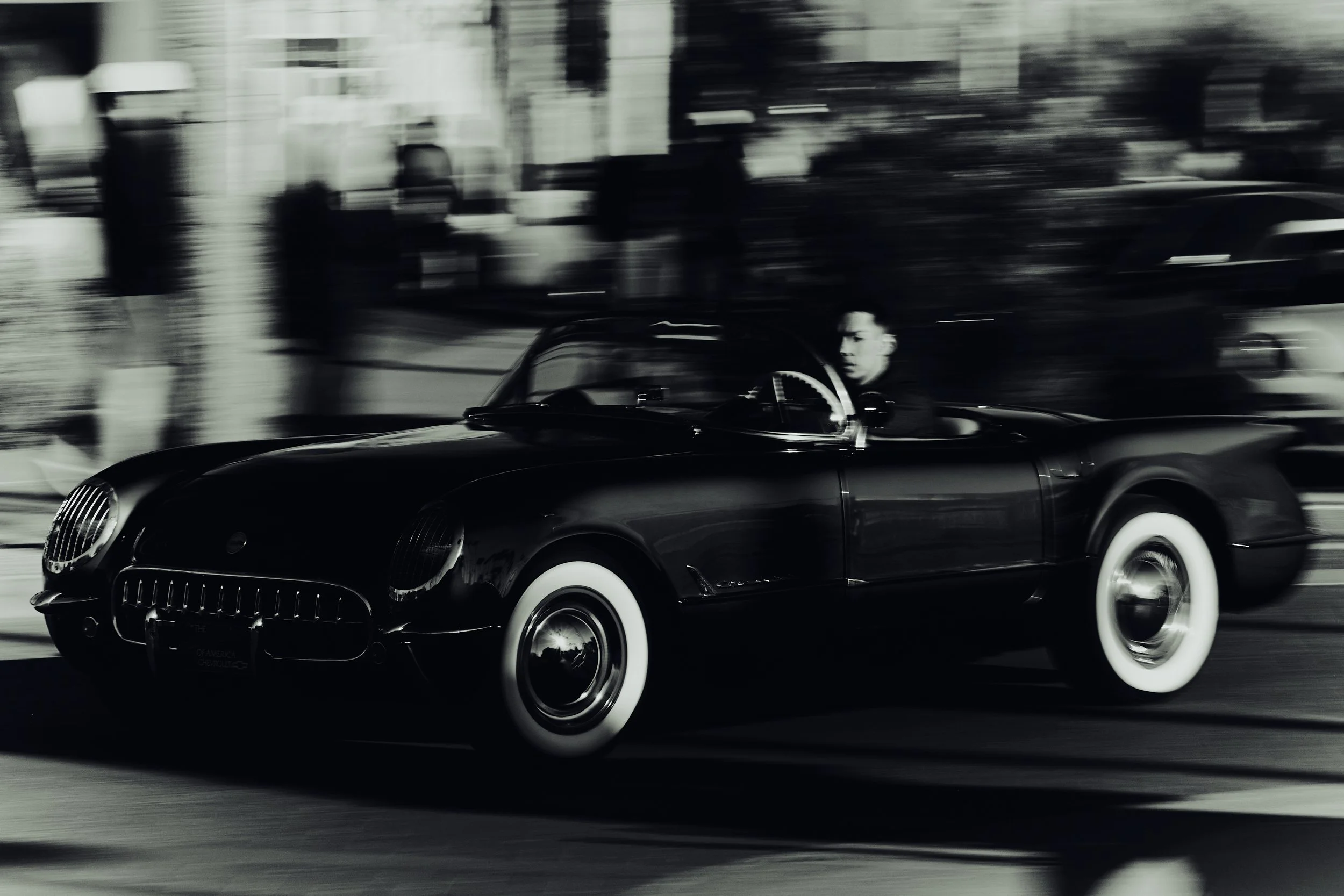 Black vintage convertible car with white-walled tires driving on a city street, with a man in the driver's seat, in black and white, motion blurred background.