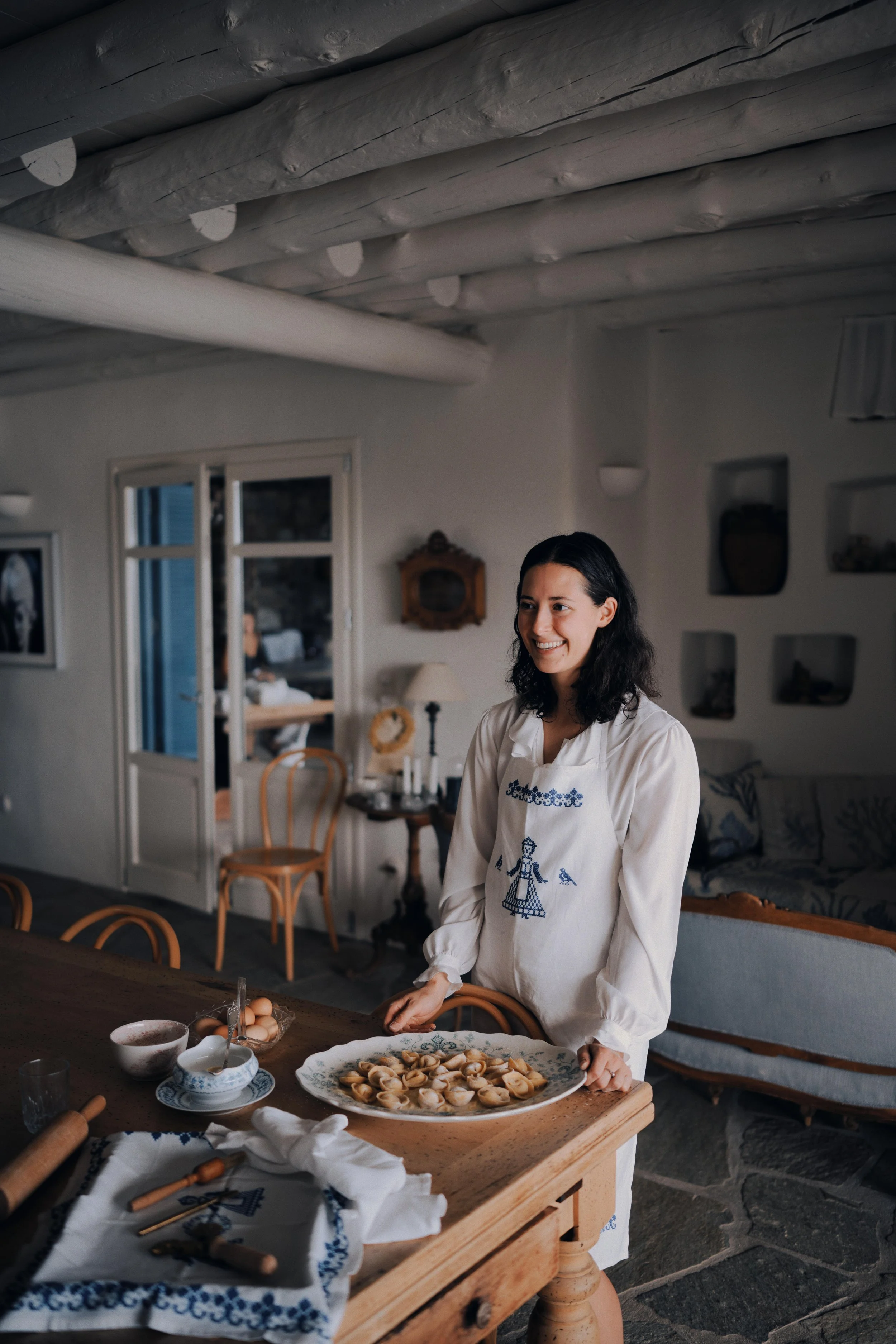 A woman standing in a cozy, rustic kitchen holding a large plate of homemade dumplings, with various cooking utensils and ingredients on the wooden table in front of her.