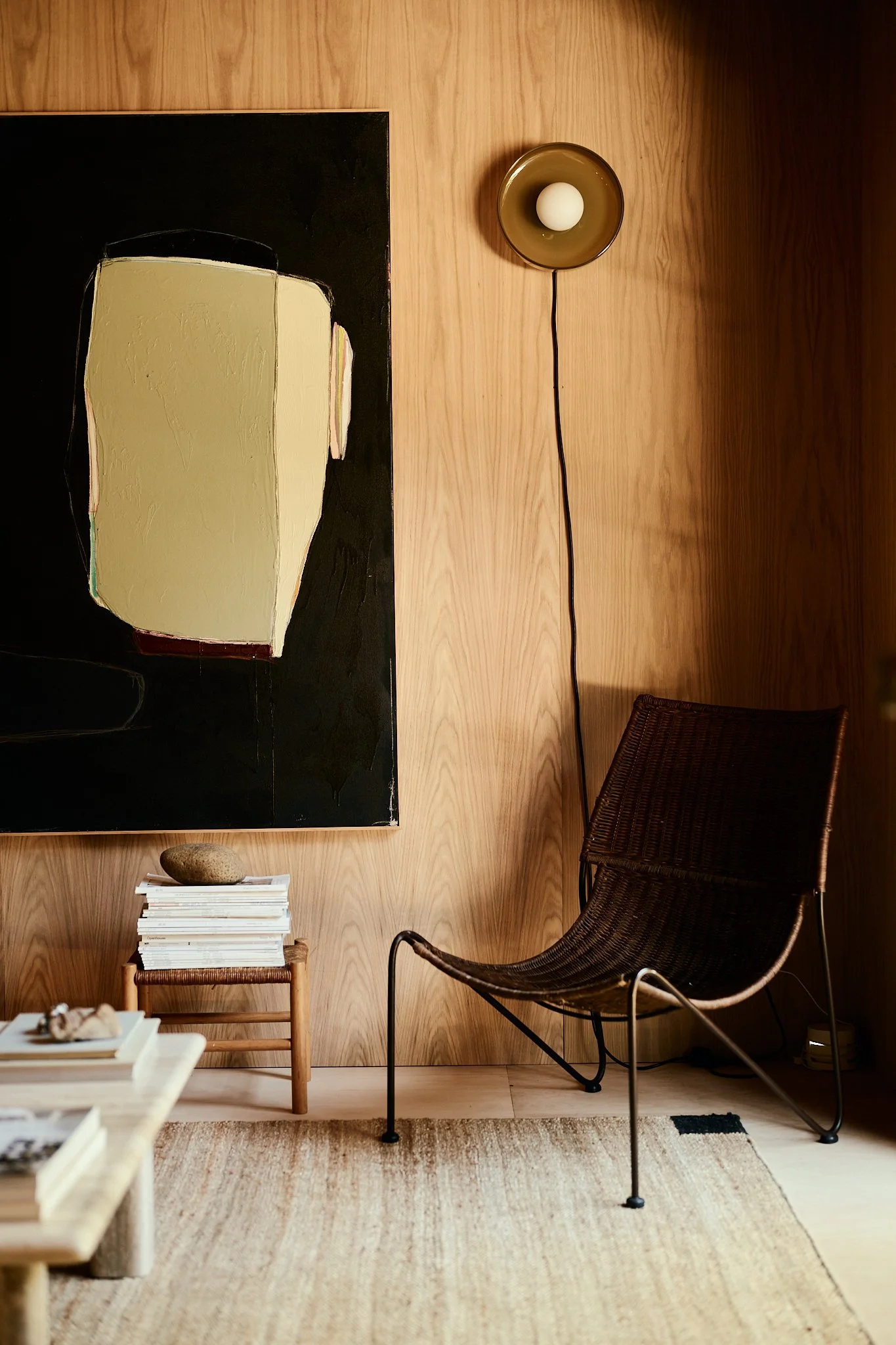 A contemporary living room corner with a black framed abstract art piece, a rattan chair, a small wooden stool with stacked books and a stone on top, a beige rug, and a round wall-mounted lamp with an egg-shaped bulb against a wood-paneled wall.