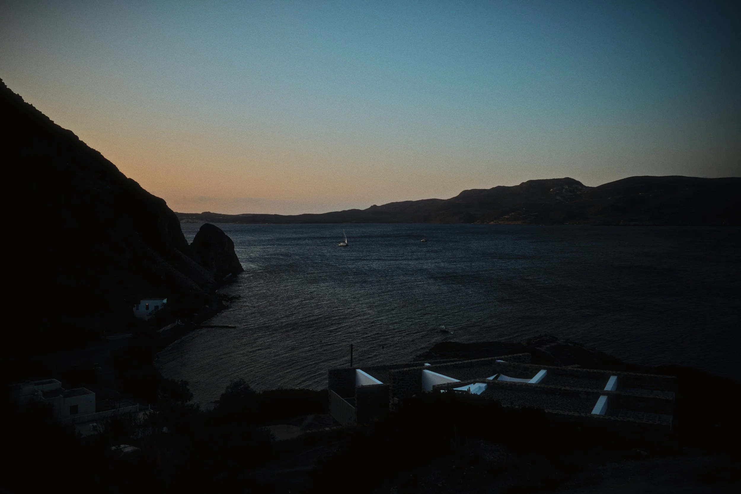 A coastal scene at dusk with a bay, sailboats, boats in the water, and hills in the background. The foreground shows dark buildings and hills on the left side.