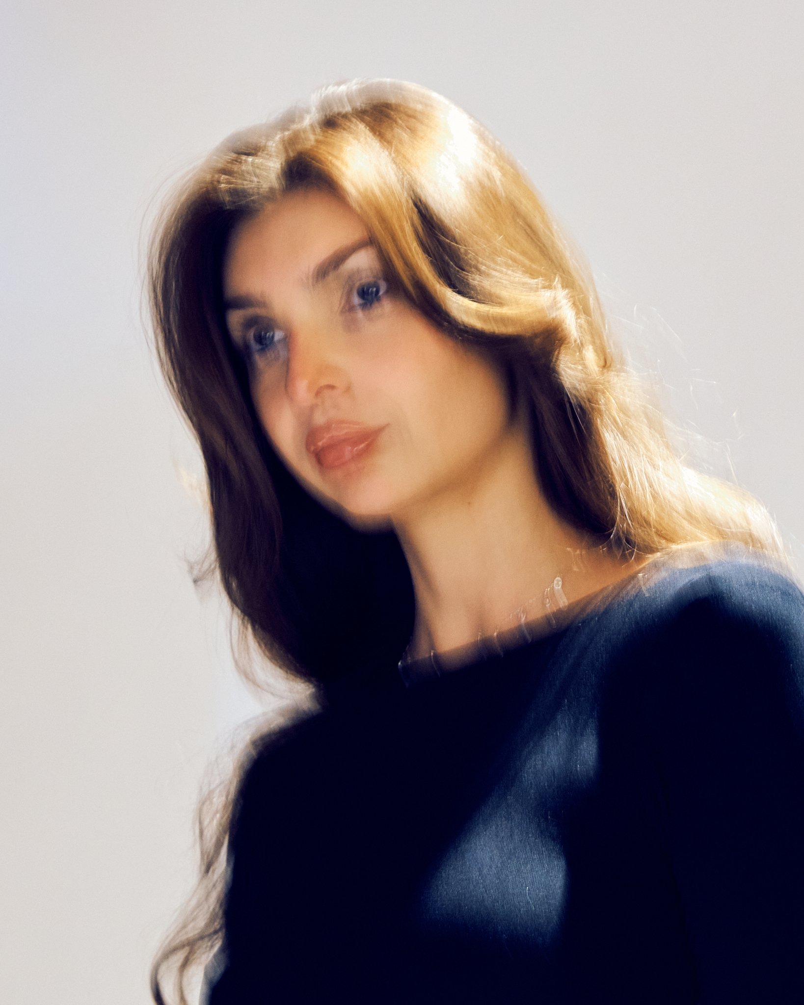 A woman with long, wavy, brown hair with highlights, wearing a black top, looking slightly to the side with a soft expression.