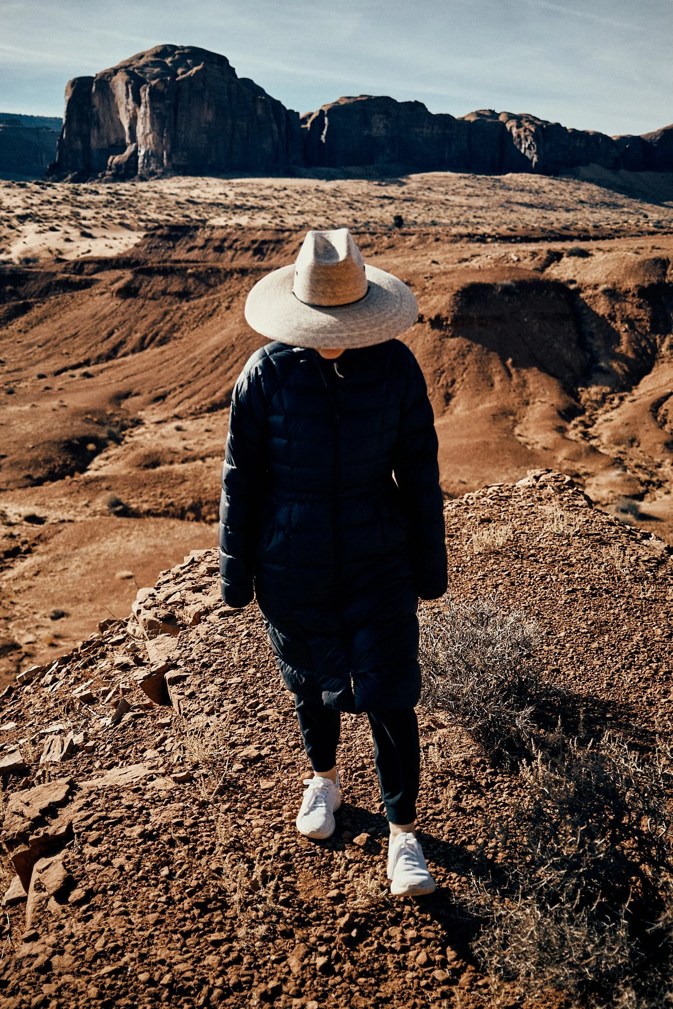 Person walking on rocky trail in desert landscape with large rock formations in background, wearing a wide-brimmed hat, dark padded coat, and white sneakers.