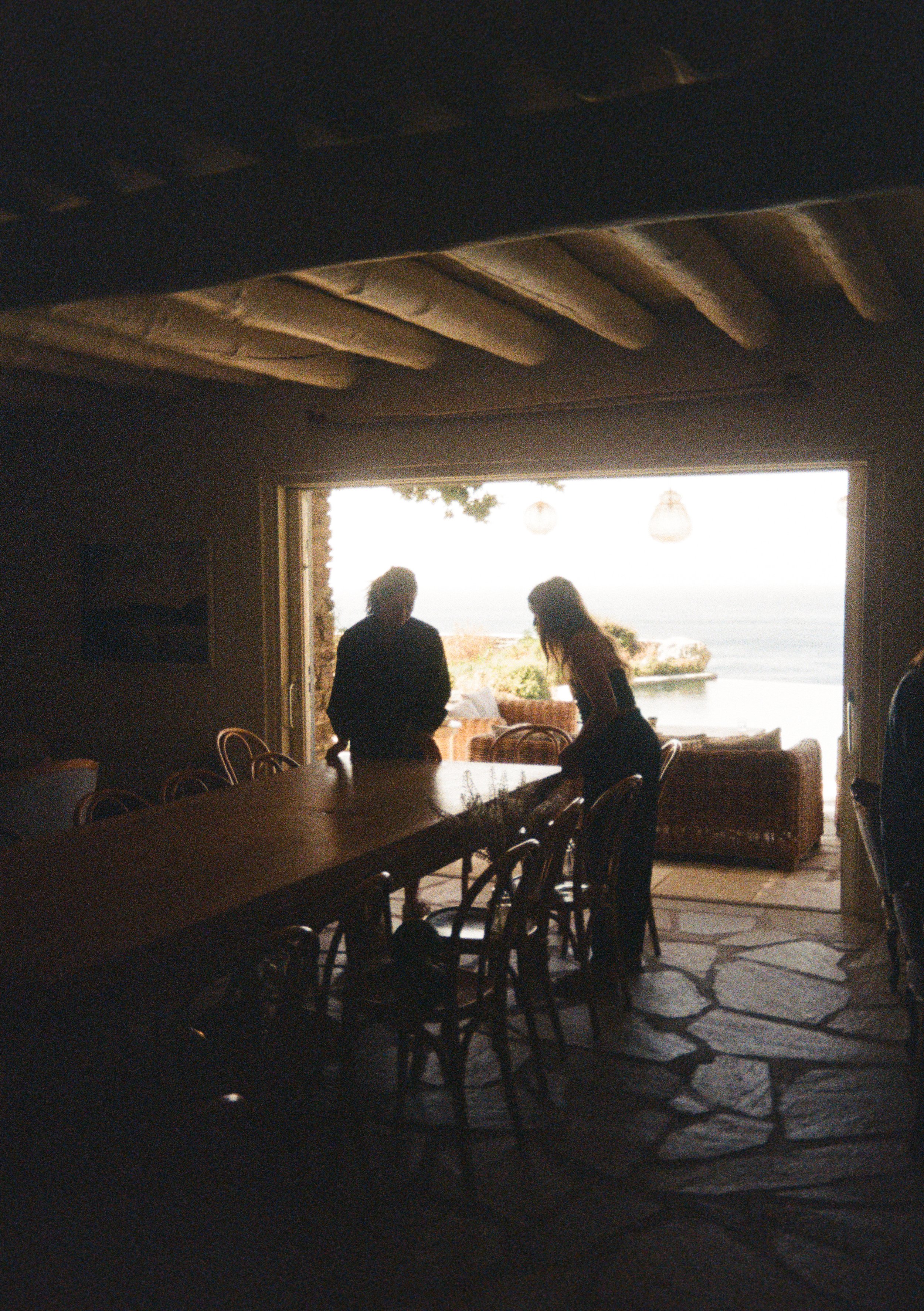 Two people silhouetted inside a room near a large open window, overlooking the ocean, with outdoor seating and potted plants visible.
