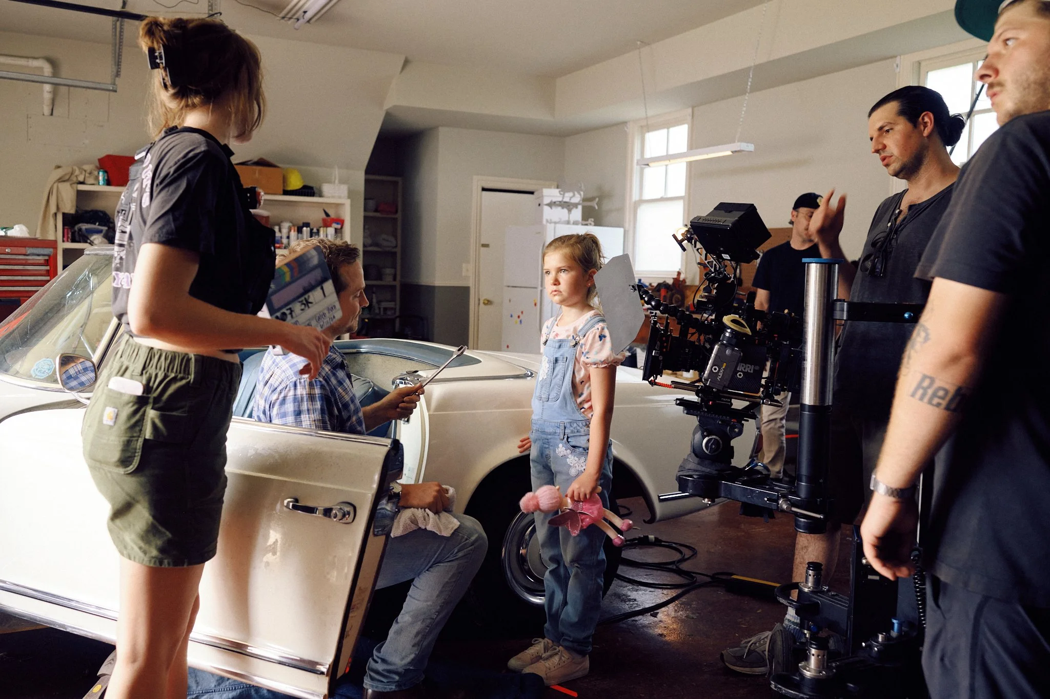 A film crew filming a scene with a young girl standing next to a vintage white car in a garage setting.