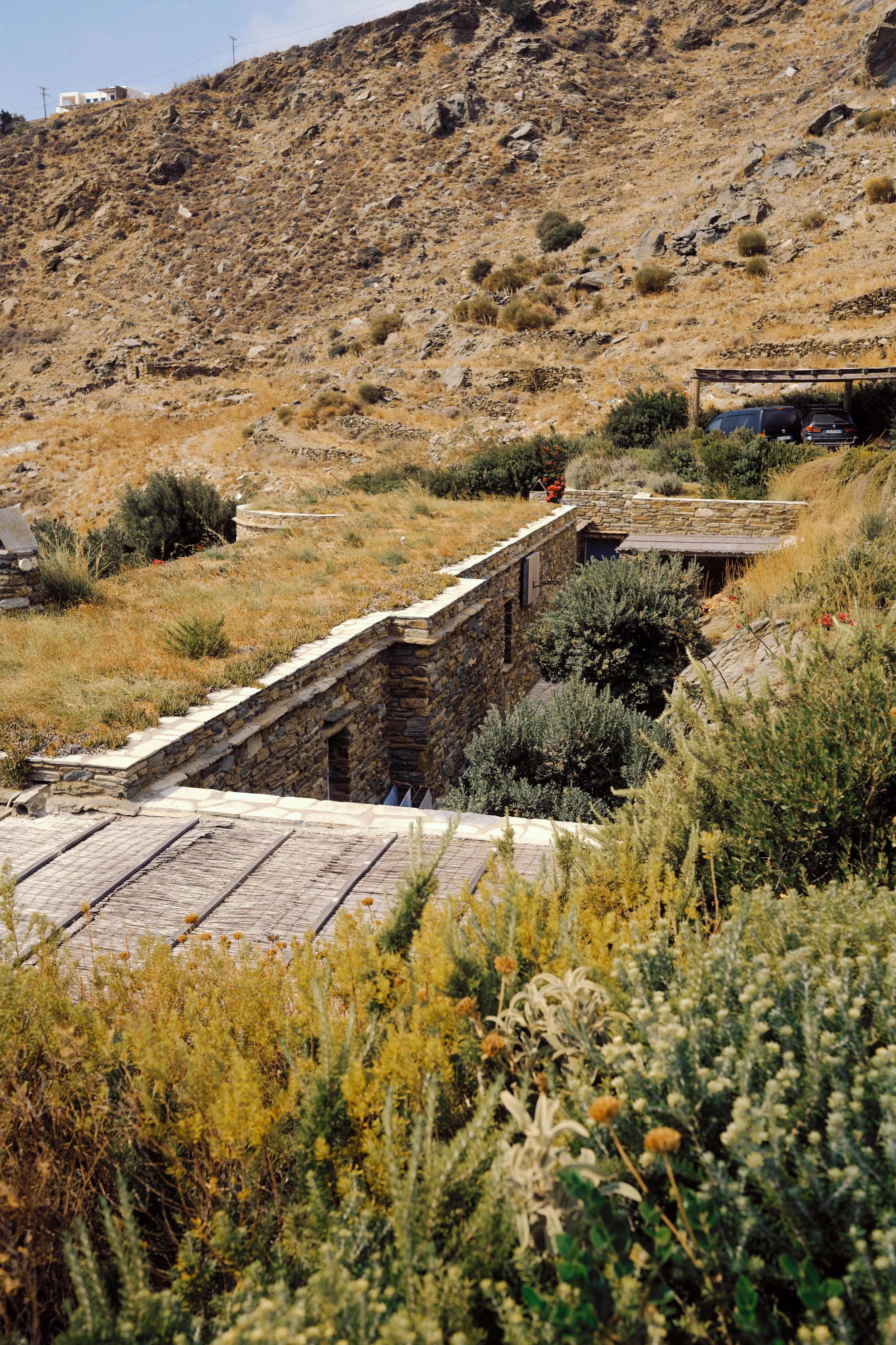 A rustic stone house built into a hillside with a grassy roof, surrounded by desert-like terrain and vegetation, with a parking area and hills in the background.