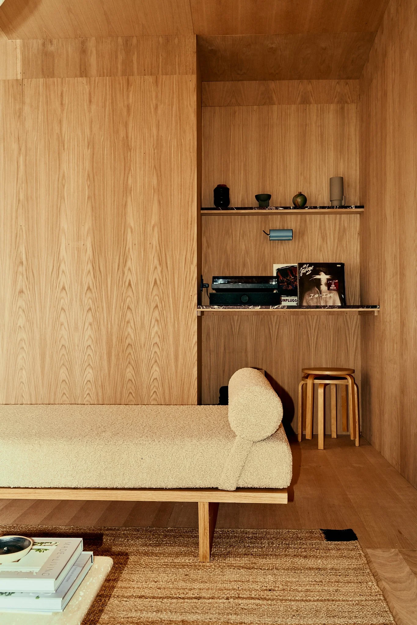 A minimalist living room corner with a beige textured sofa, a stack of books on a small table, a woven rug, wooden stools, and built-in wooden shelves displaying decorative objects and records.