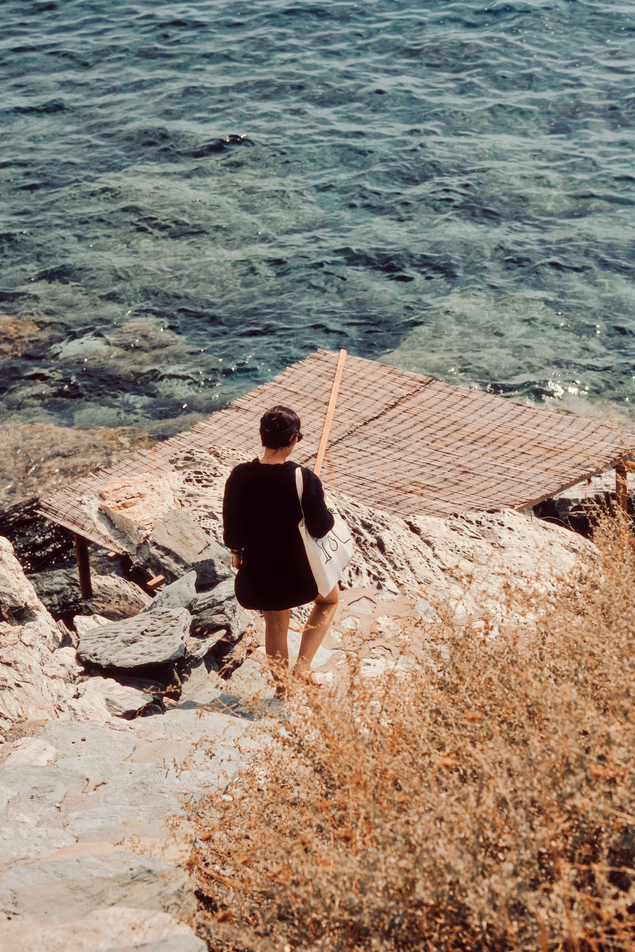 A woman in black clothing carrying a white tote bag, walking down stone steps towards the water at a rocky seaside, with a wooden platform and clear blue water in the background.
