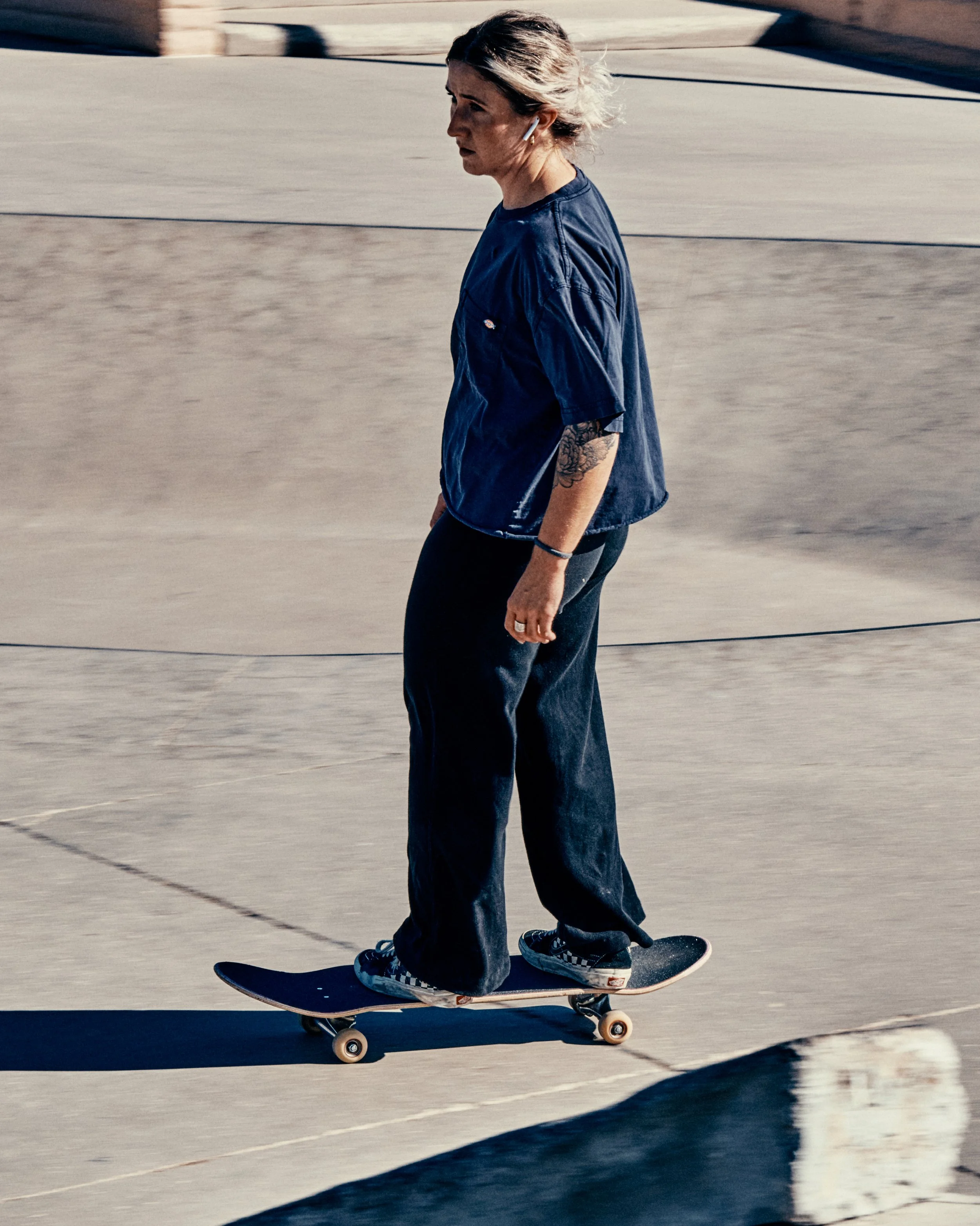 A woman skateboarding on a concrete surface, wearing a blue T-shirt, black pants, and checkered sneakers, with earbuds in her ears and tattoos on her left arm.