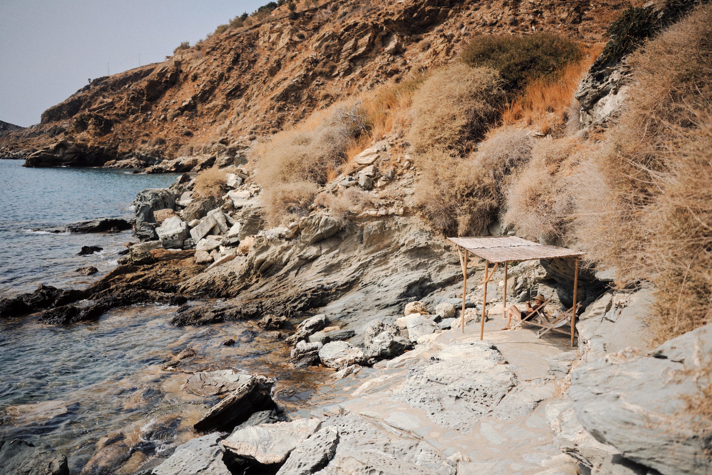 A woman relaxing on a lounge chair under a small canopy built from wooden sticks and a fabric roof, near rocky shoreline, with dry bushes and a hillside in the background.