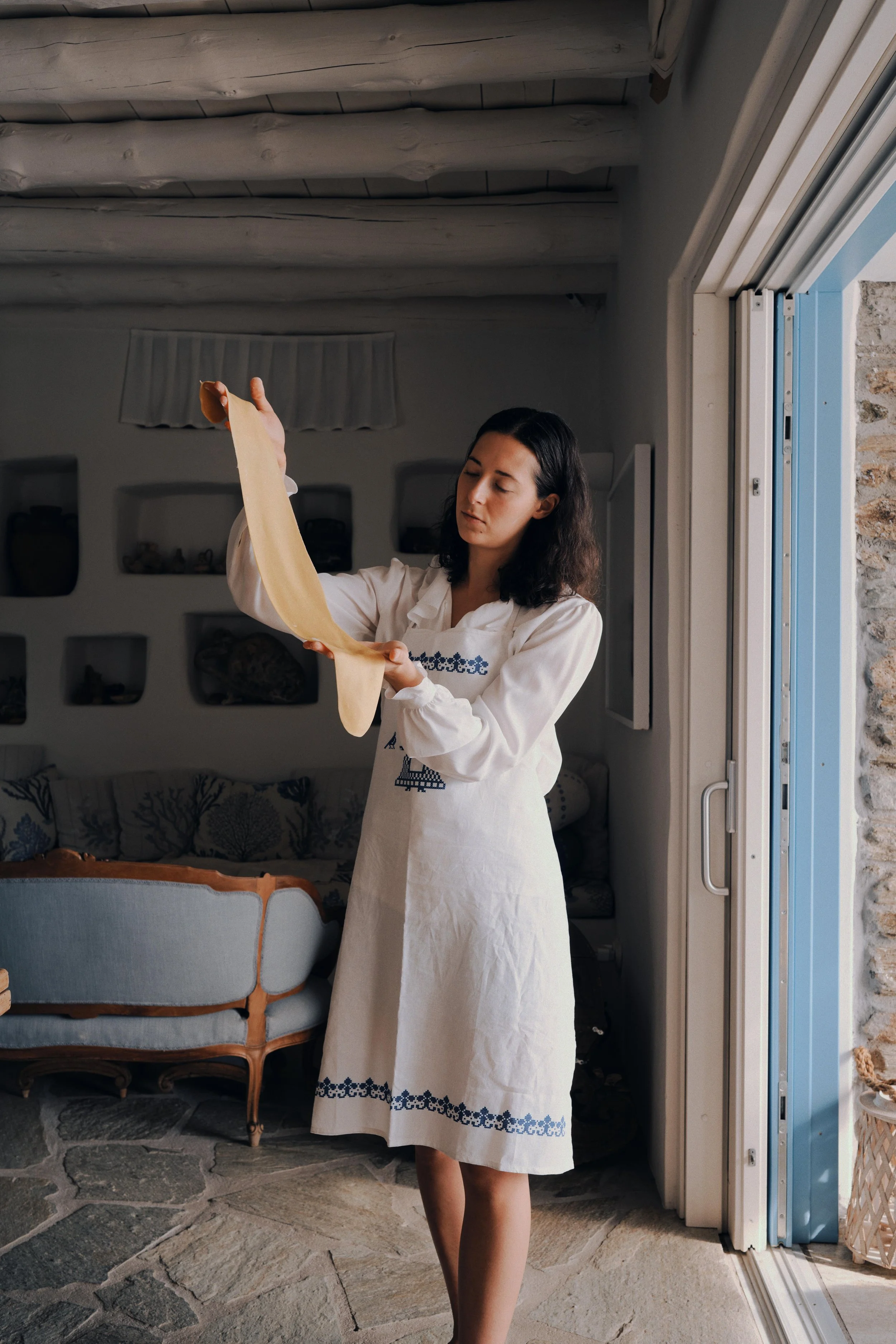 A woman in a white embroidered dress is stretching and preparing to bake bread in a cozy home kitchen with stone flooring, a blue door, and a small sofa in the background.