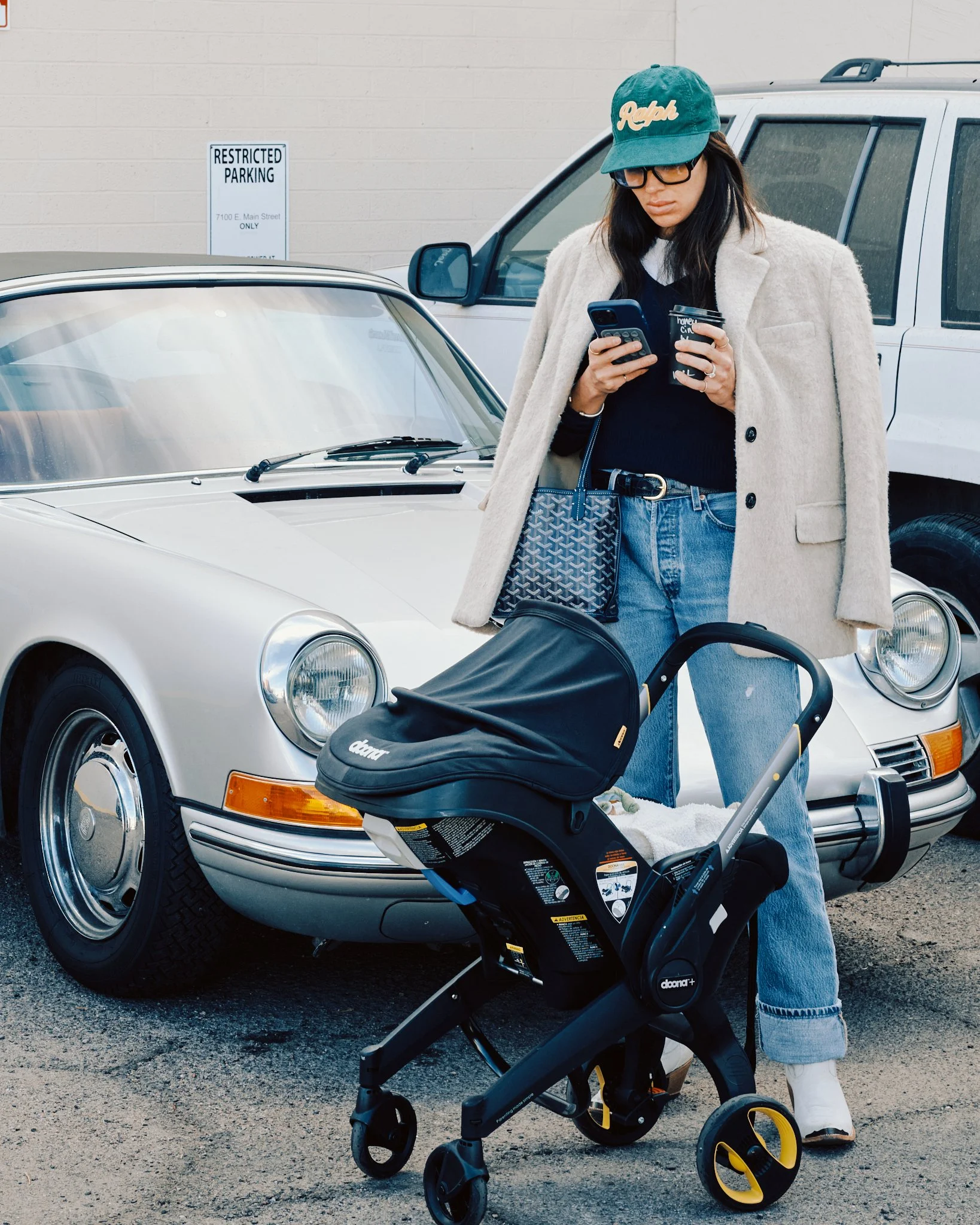 A woman standing in a parking lot, looking at her phone, with a baby stroller, a purse, and two cars, one vintage and one modern, in the background.